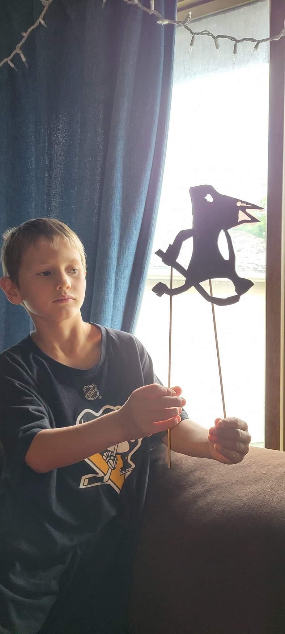 A young boy sitting indoors, holding a stick with a paper cutout of a robot figure on it, near a window with blue curtains and string lights.