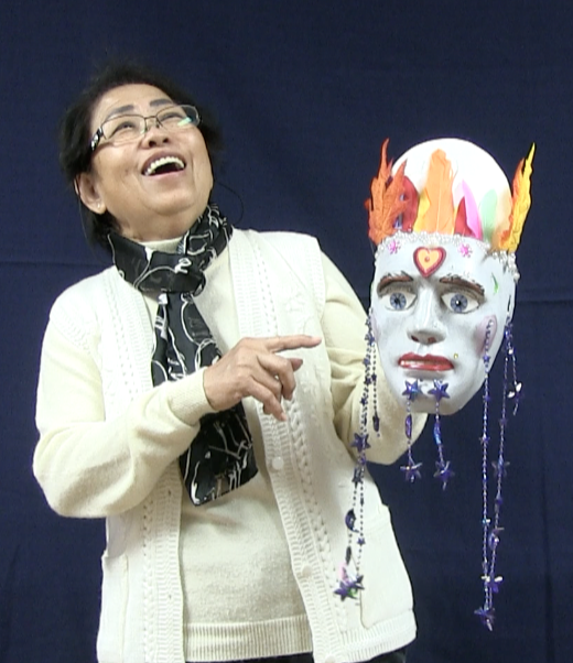 Asian woman smiles as she holds up a mask- puppet