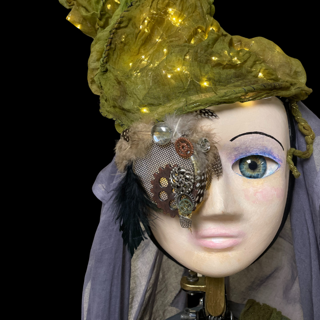 A puppet with a human face, and light sparkling green head dress and an augmented eyepiece
