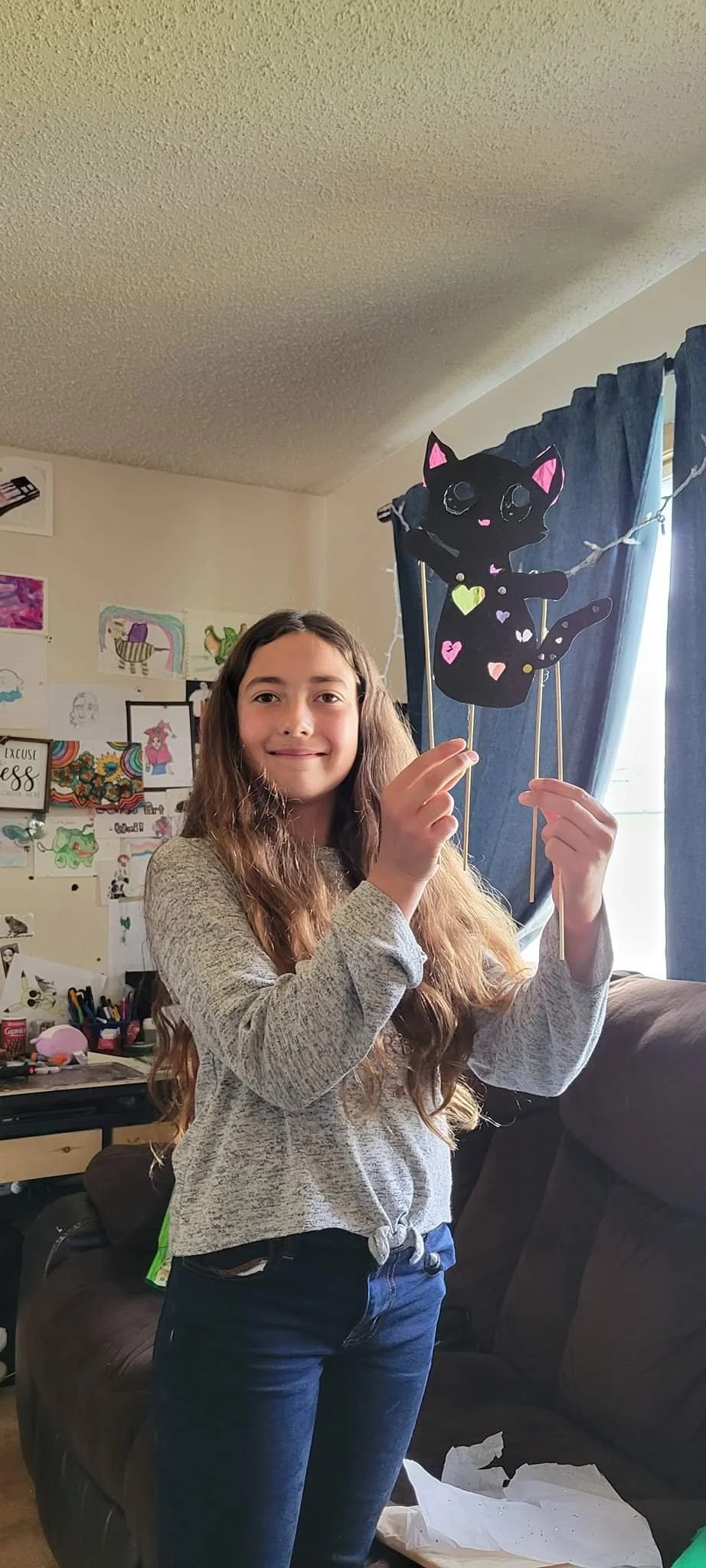 A girl with long curly hair smiling and holding a black cat-shaped puppet with colorful heart-shaped decorations, standing inside a room with drawings on the wall and blue curtains in the background.