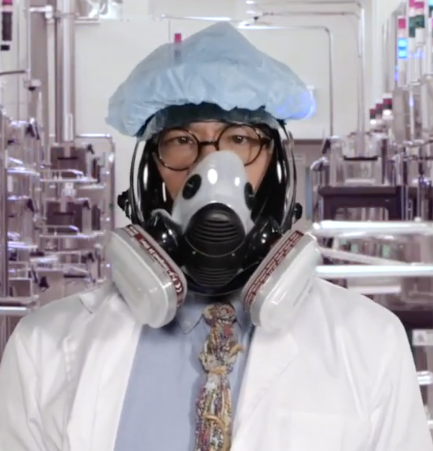 Person wearing a gas mask and blue shower cap in front of a lab scene 