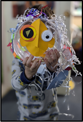 Child holding a colorful handmade craft resembling an owl with large eyes and a beak, made from paper, plastic, and decorative streamers.