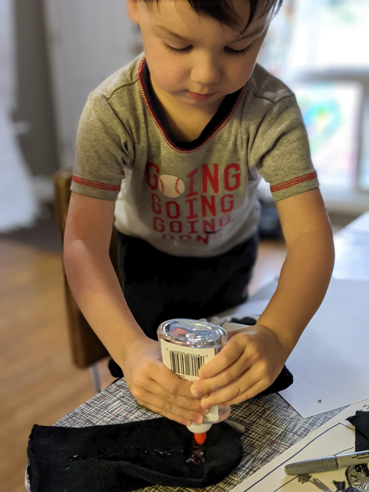 A young boy in a gray T-shirt with red lettering is squeezing glue from a bottle onto a piece of black fabric on a table.
