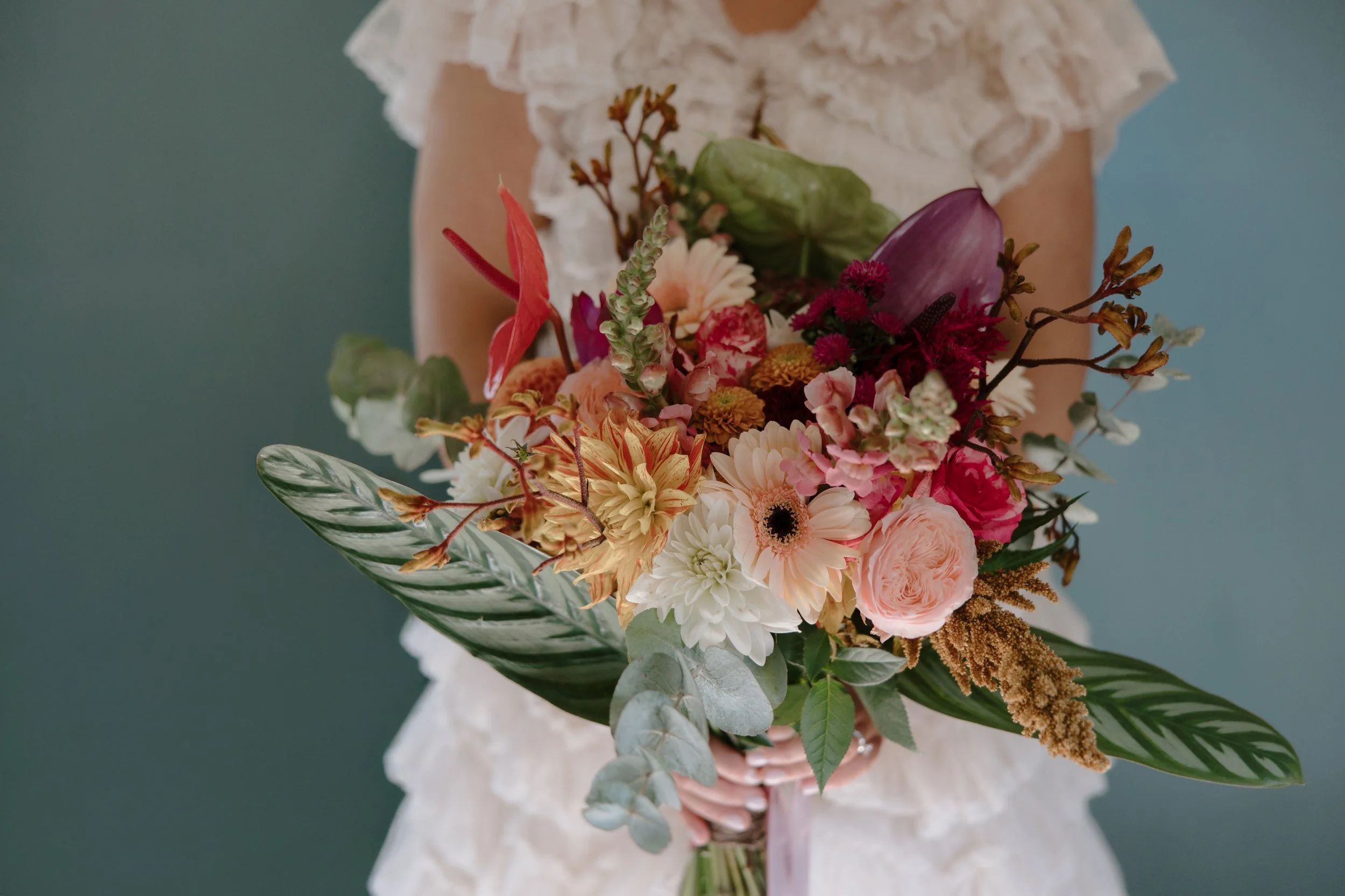 Person holding a colorful bouquet of flowers, including roses, dahlias, anthuriums, and various greenery, against a muted teal background.