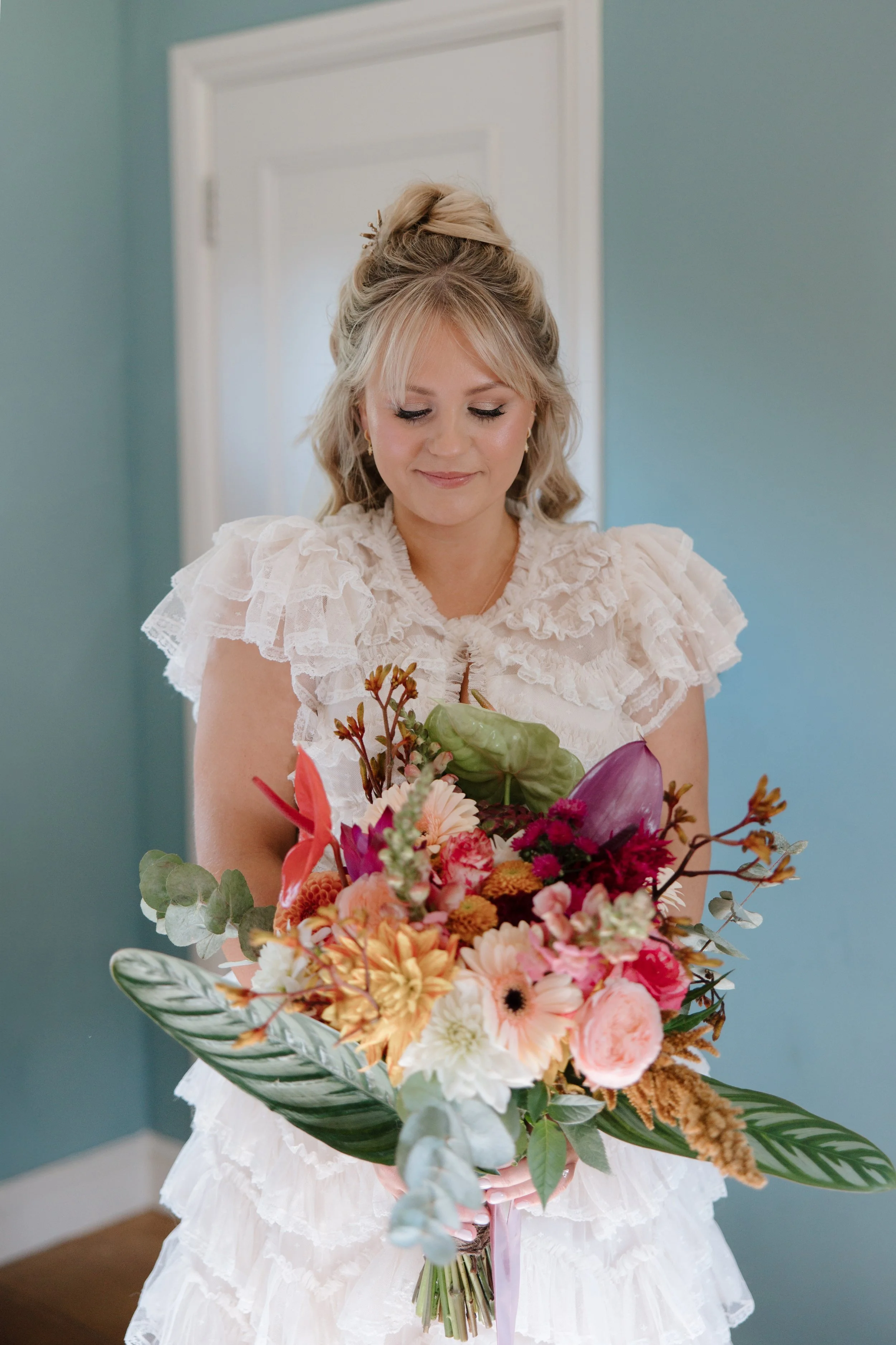 A woman in a white ruffled dress holding a colorful bouquet of flowers, standing in a room with a blue wall and white door in the background.