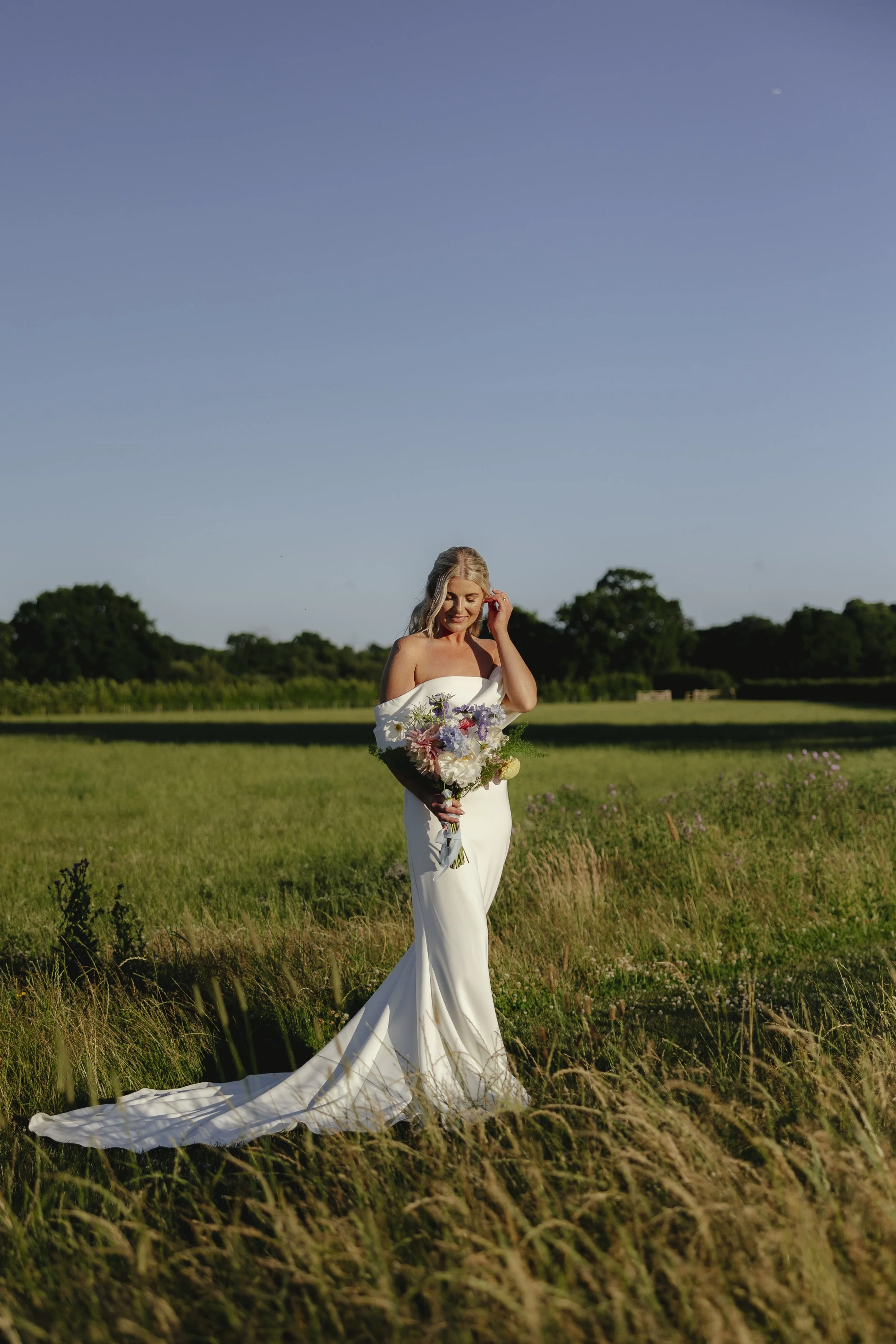 A woman in a white wedding dress standing in a grassy field holding a bouquet of purple, pink, and white flowers, with trees and a clear blue sky in the background.