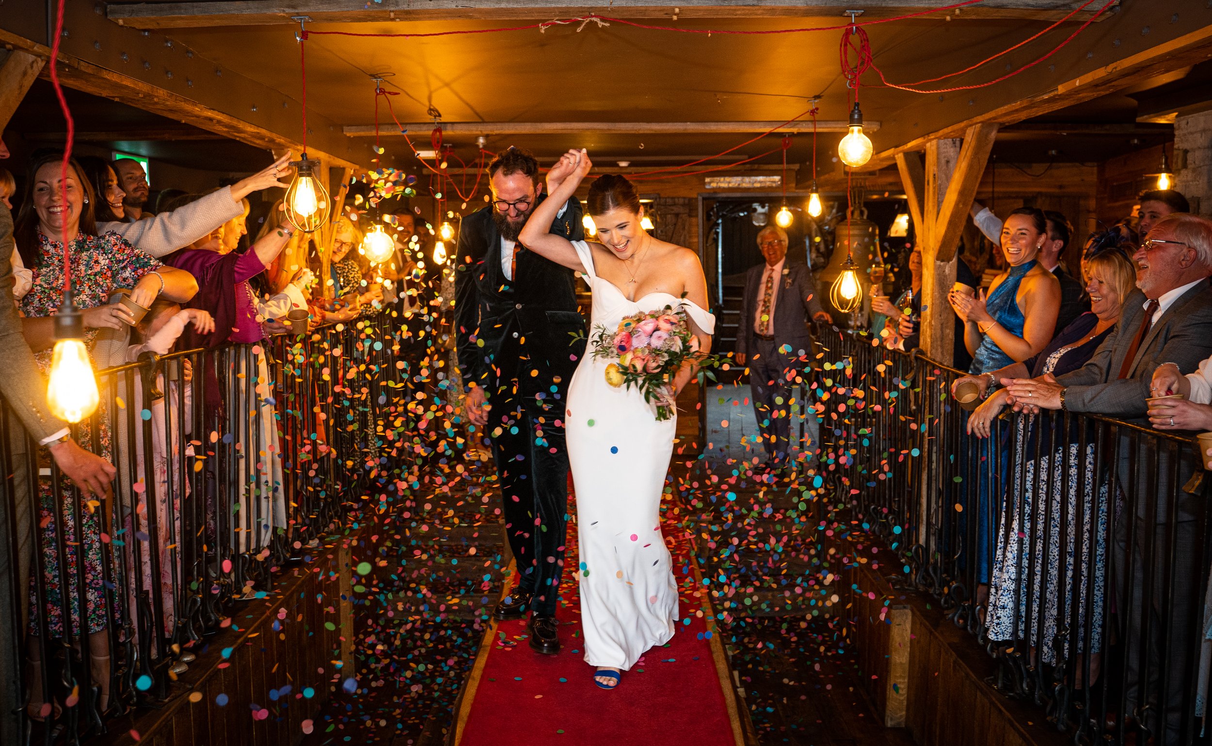 Bride and groom walking through a confetti-filled aisle, smiling, surrounded by cheering guests in a rustic venue with hanging lights.