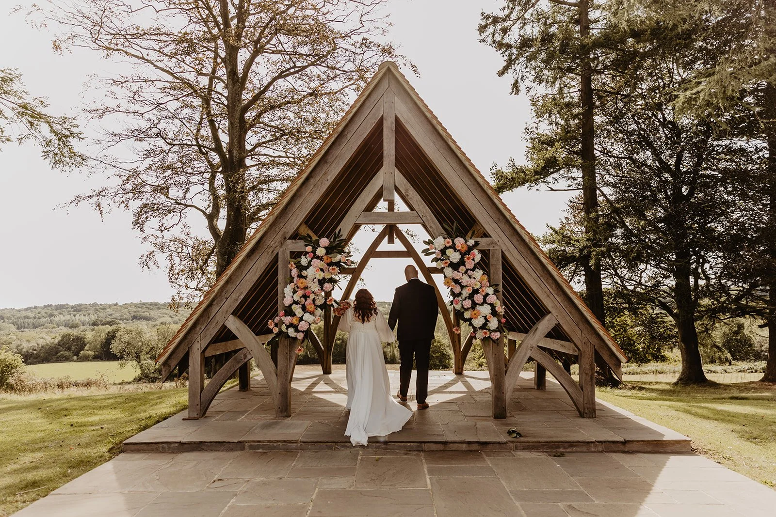 A bride and groom holding hands, walking toward an outdoor wedding ceremony Pavilion decorated with pink, white, and peach flowers.
