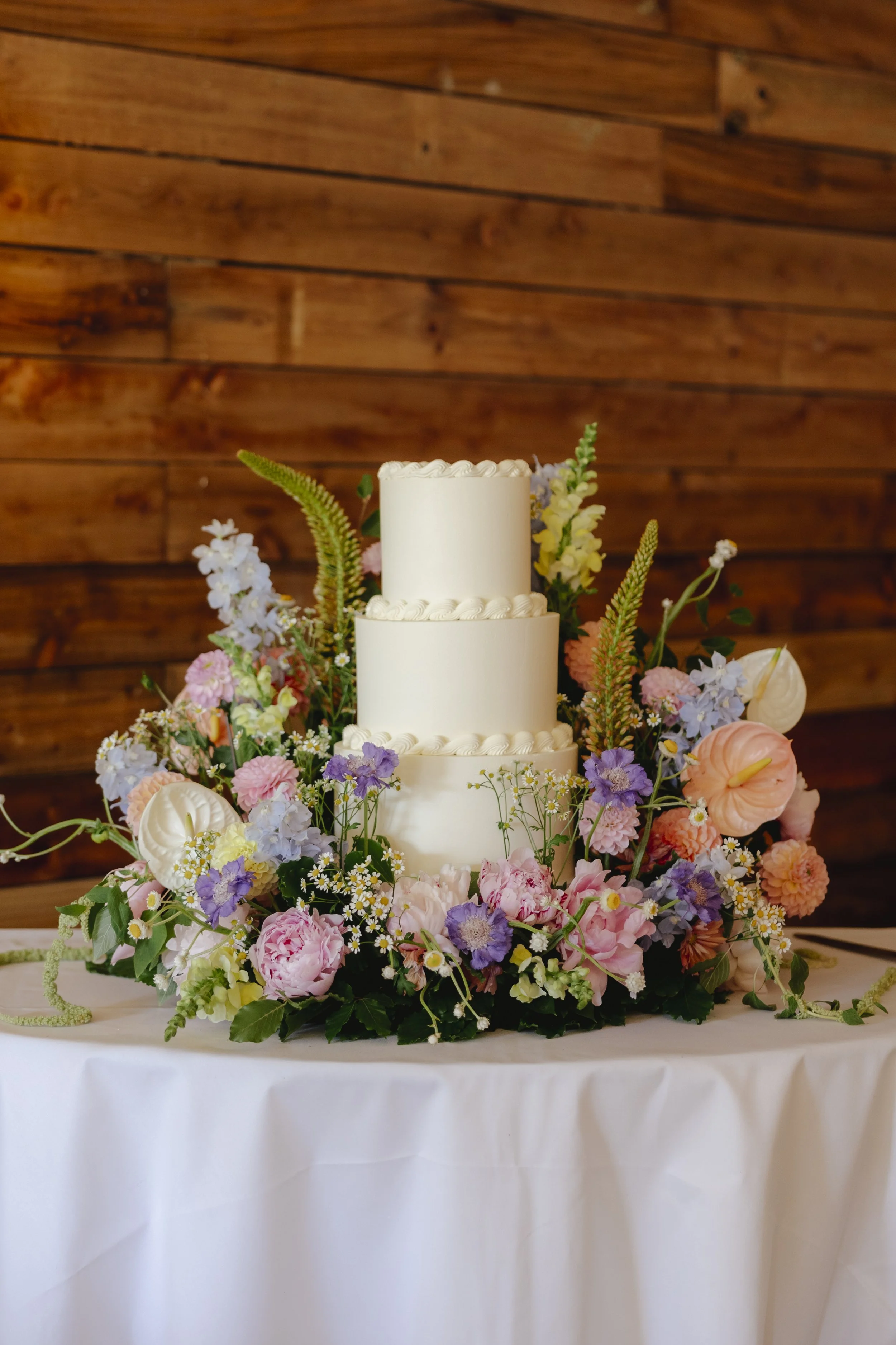 Three-tier white wedding cake with piped decorations, surrounded by a floral arrangement with pink, purple, white, and green flowers on a table with a white tablecloth, against a wooden wall background.