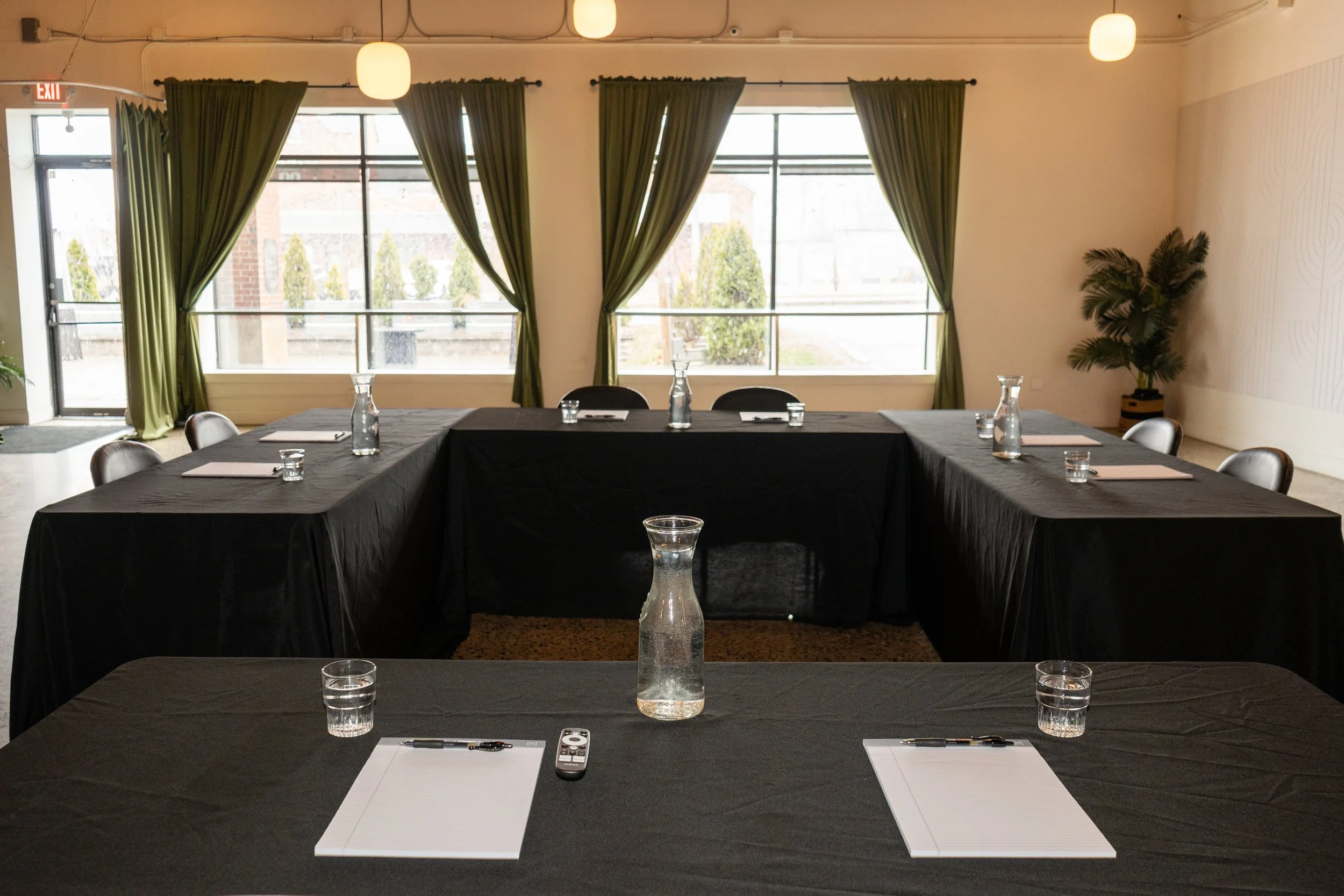 Conference room with U-shaped black table setup, notebooks, pens, water glasses, and glass pitchers, large windows with green curtains, potted plant in corner, and ceiling lights.