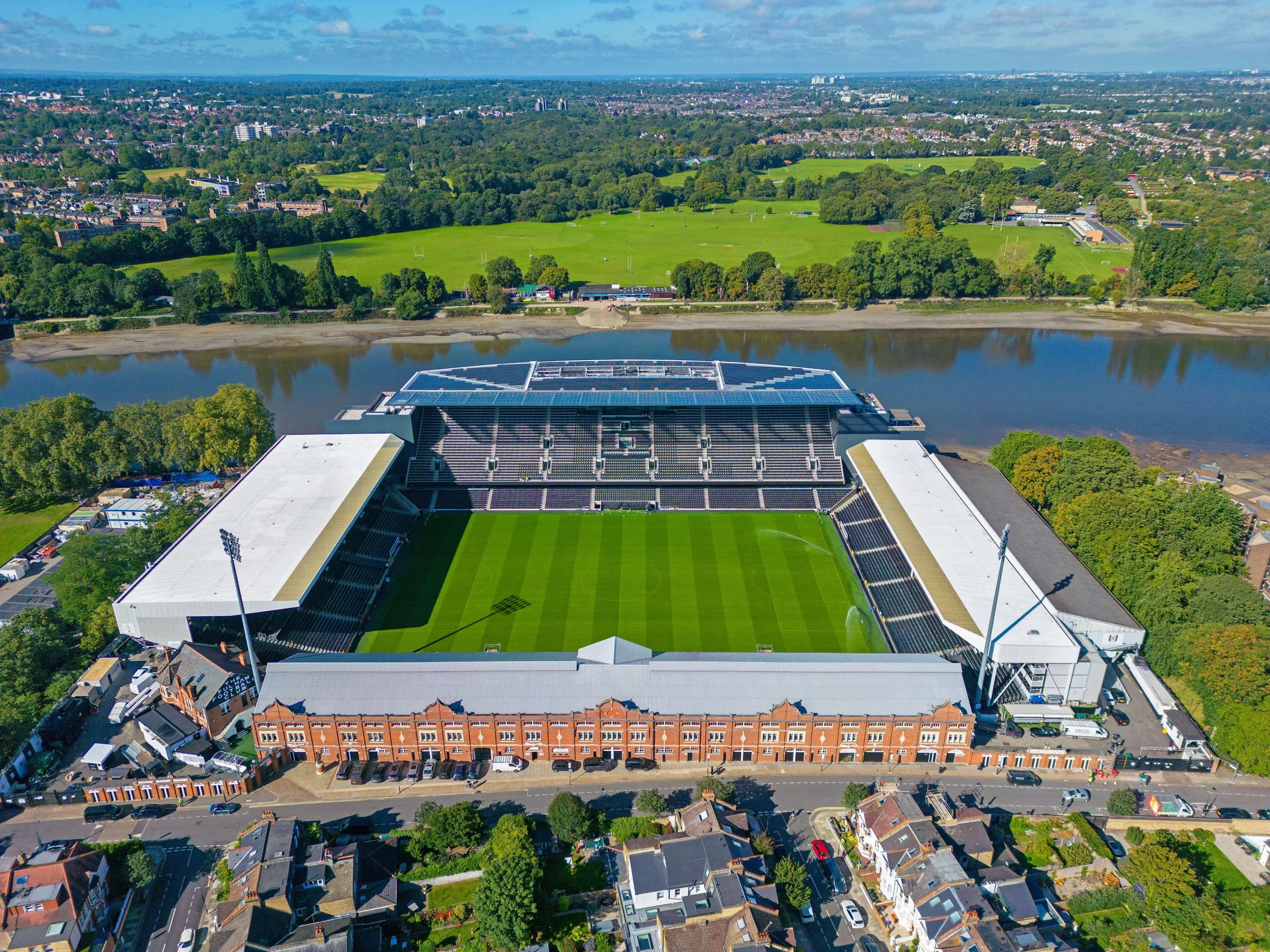An aerial view of a football stadium with green field, surrounded by a city with houses and a river in the background.