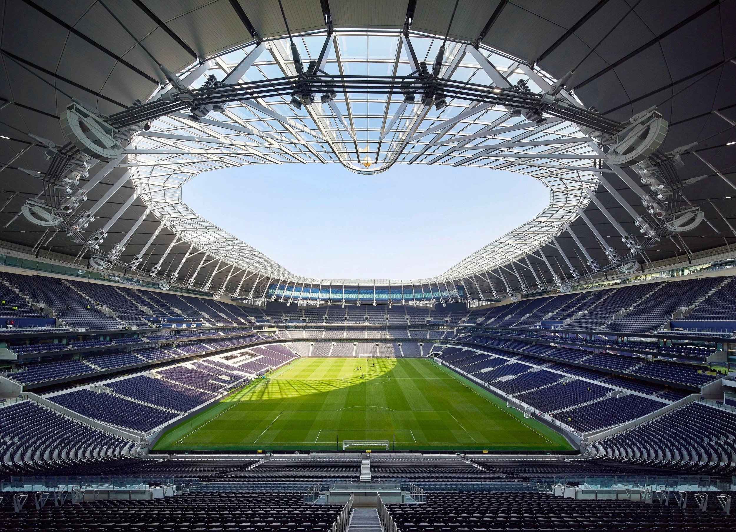 Empty modern stadium with a green soccer field, surrounded by dark blue seats, and a partially covered roof with a large open sky above.