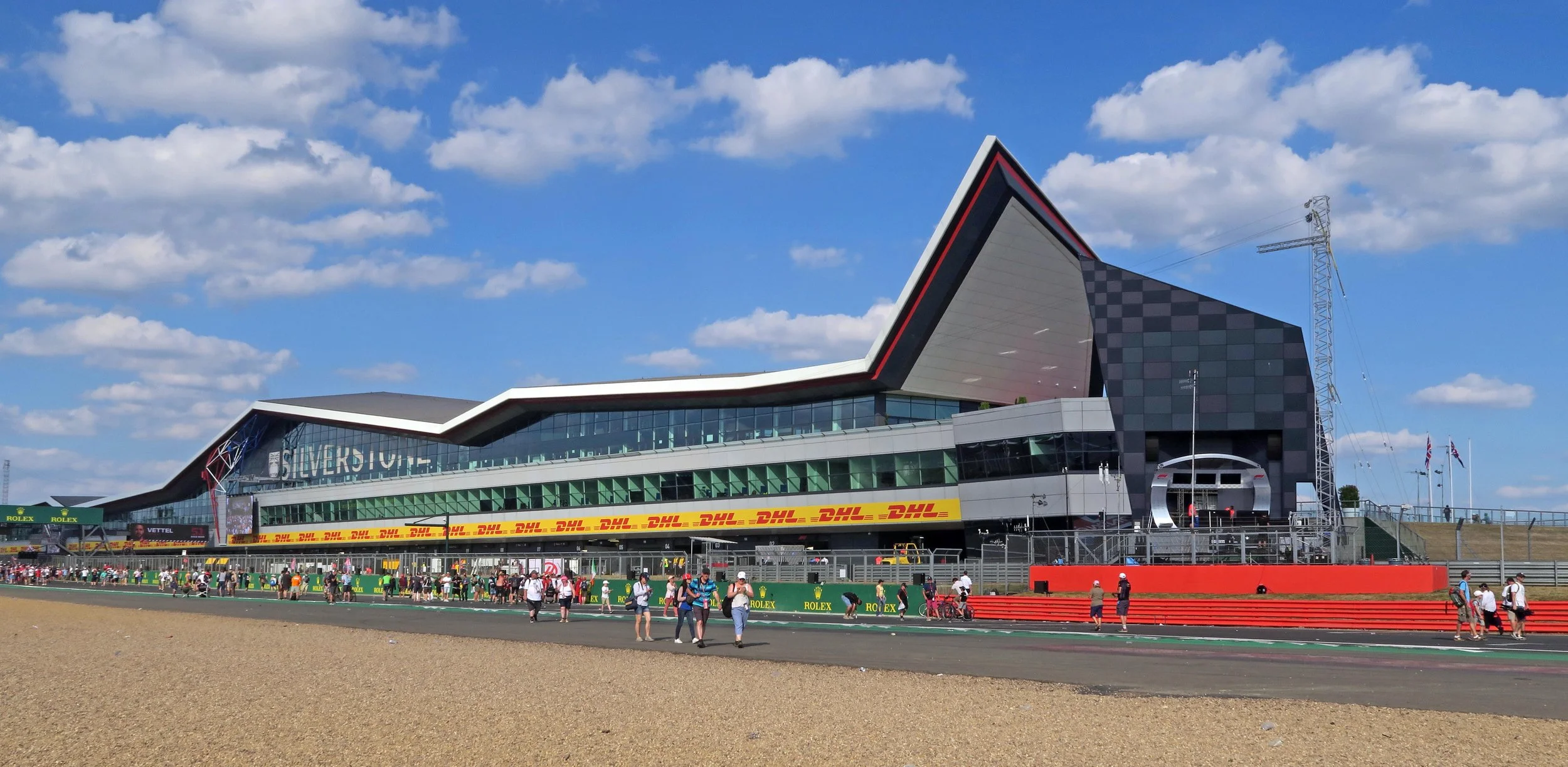 A modern racing circuit building with a distinctive angular roof, large glass windows, and banner advertisements, set against a partly cloudy sky, with people walking and gathering outside on the track area.