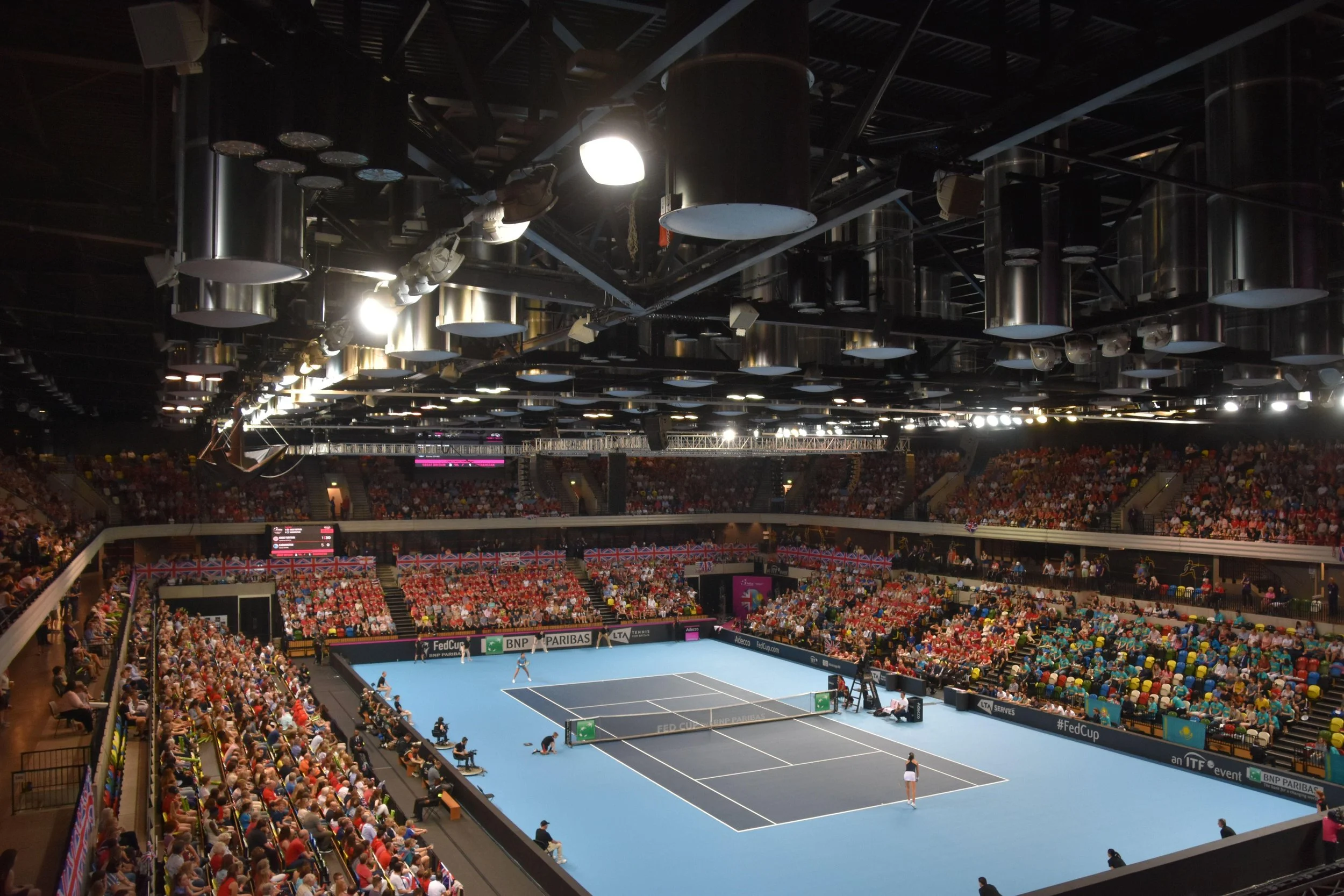 Indoor tennis stadium filled with spectators, with a blue tennis court in the center, and players preparing for a match.