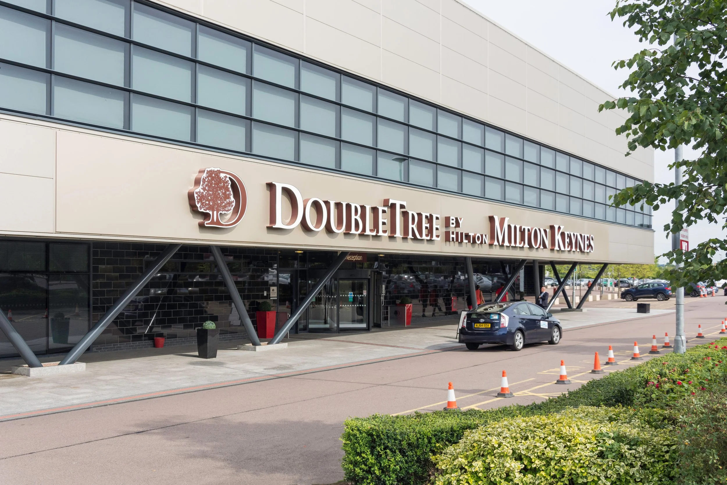 Exterior view of DoubleTree by Hilton Milton Keynes hotel, showing the entrance with large sign, glass windows, parked cars, sidewalk, and surrounding greenery.