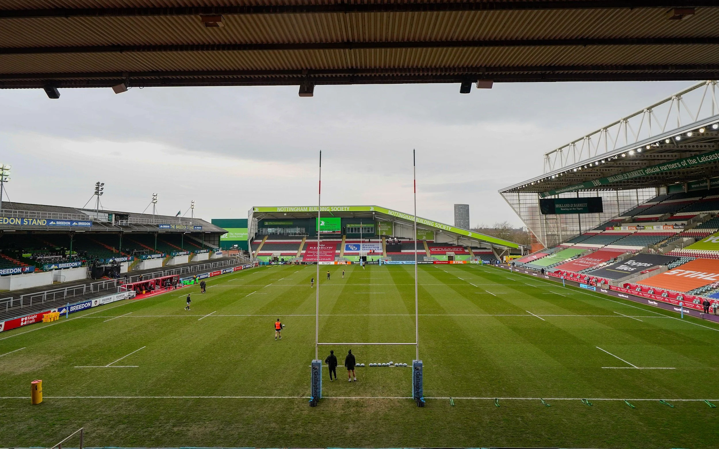 Empty rugby stadium with a well-maintained pitch and empty stands, some players and coaches practicing on the field