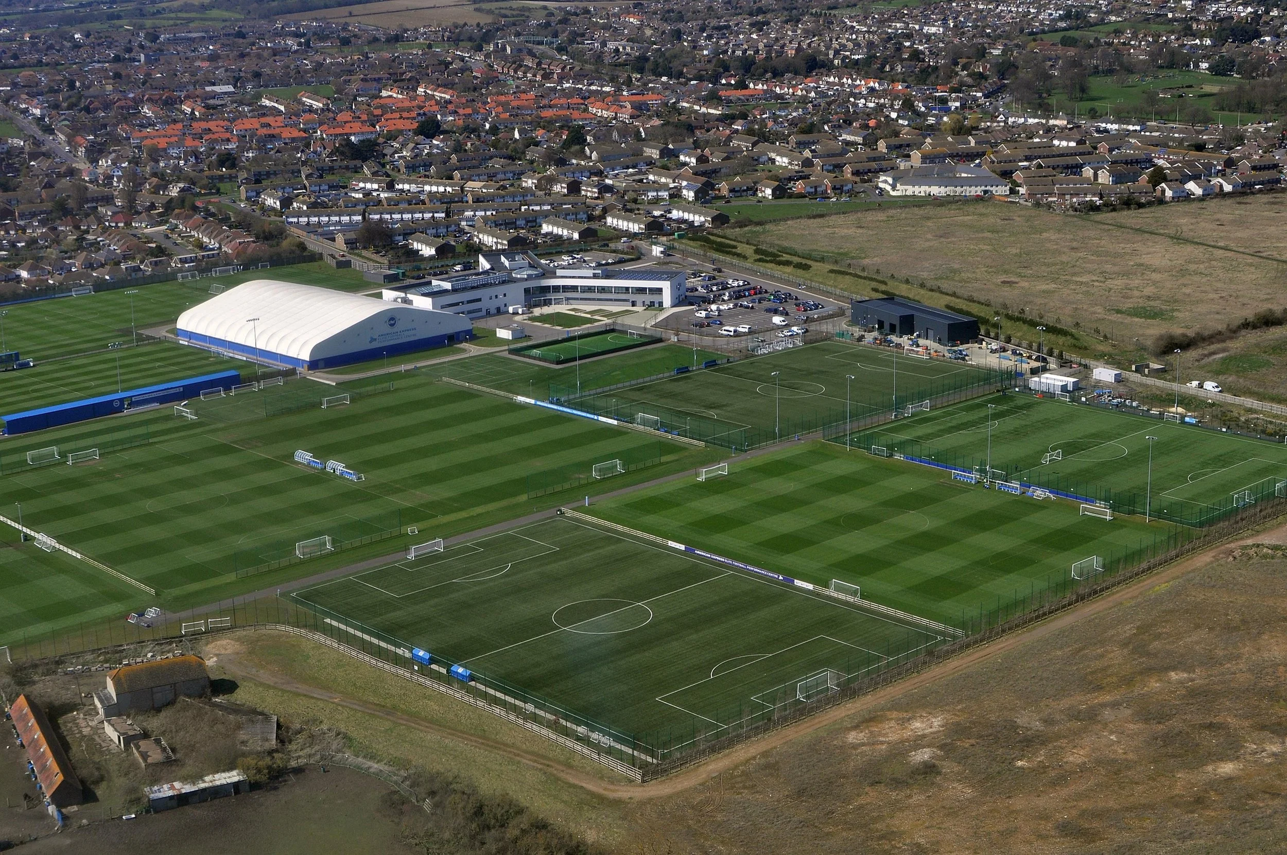 Aerial view of multiple soccer fields with green grass, goal nets, and a large white sports facility, surrounded by a parking lot and residential area.
