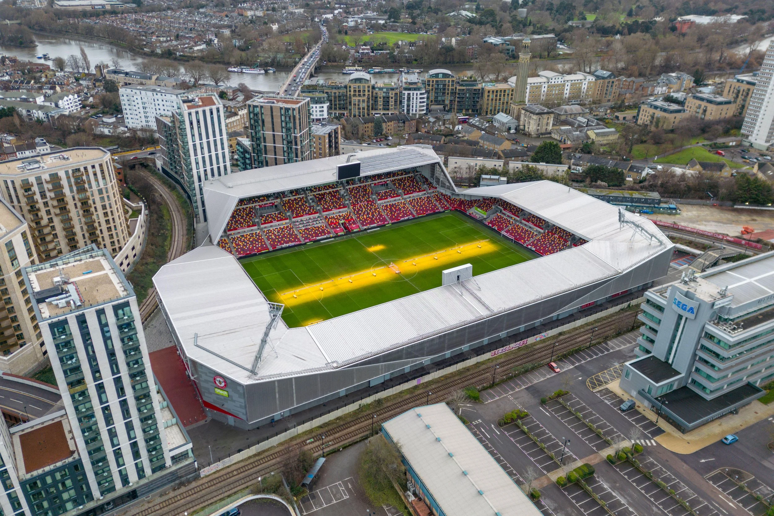 Aerial view of a football stadium surrounded by tall buildings in an urban area, with a river and bridges in the background. The stadium has a bright green field with lighting, red and yellow seating, and a large screen on one side.