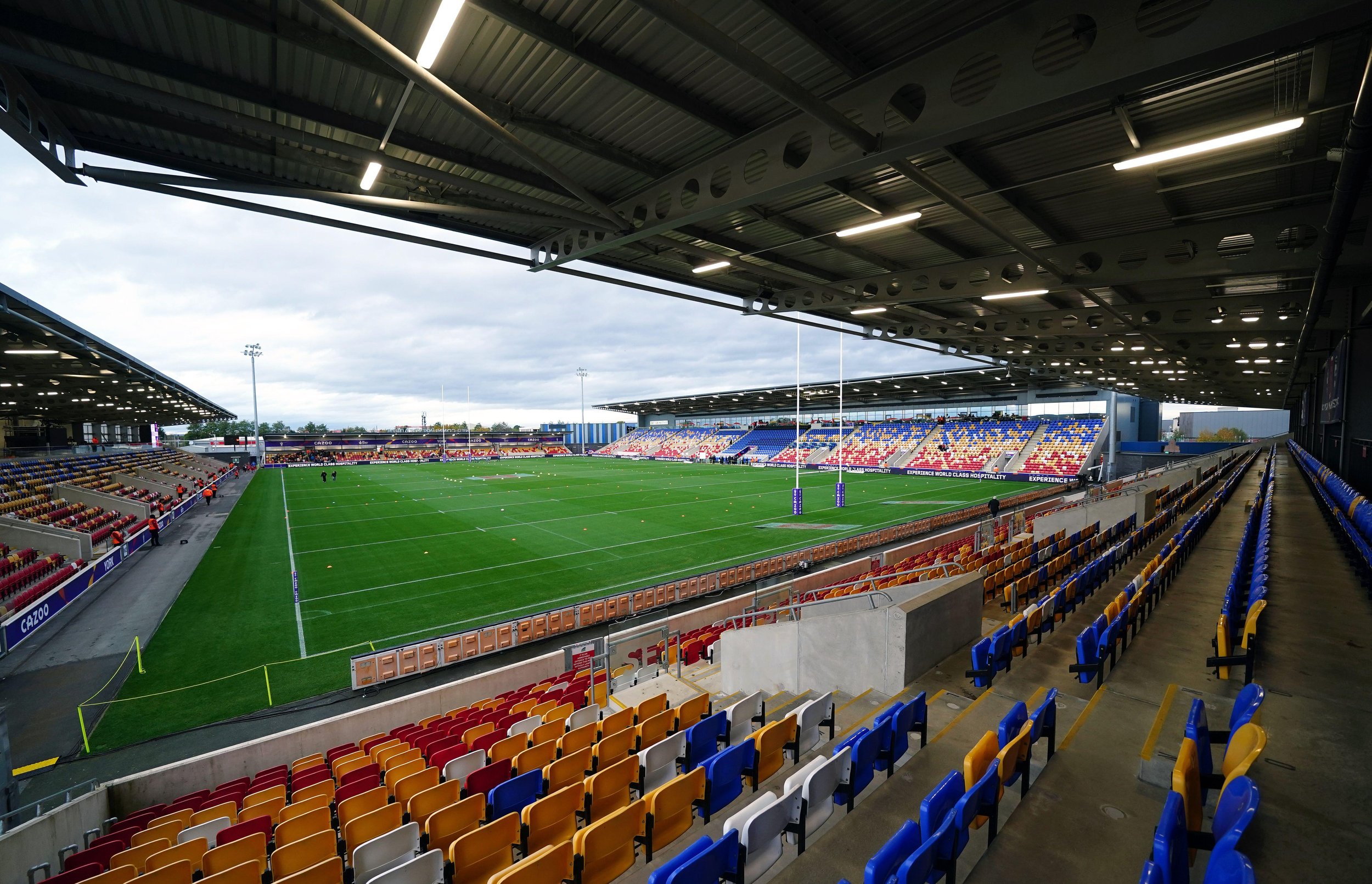 Empty rugby stadium with colorful seats, a well-maintained green field, goal posts, and an overcast sky overhead.
