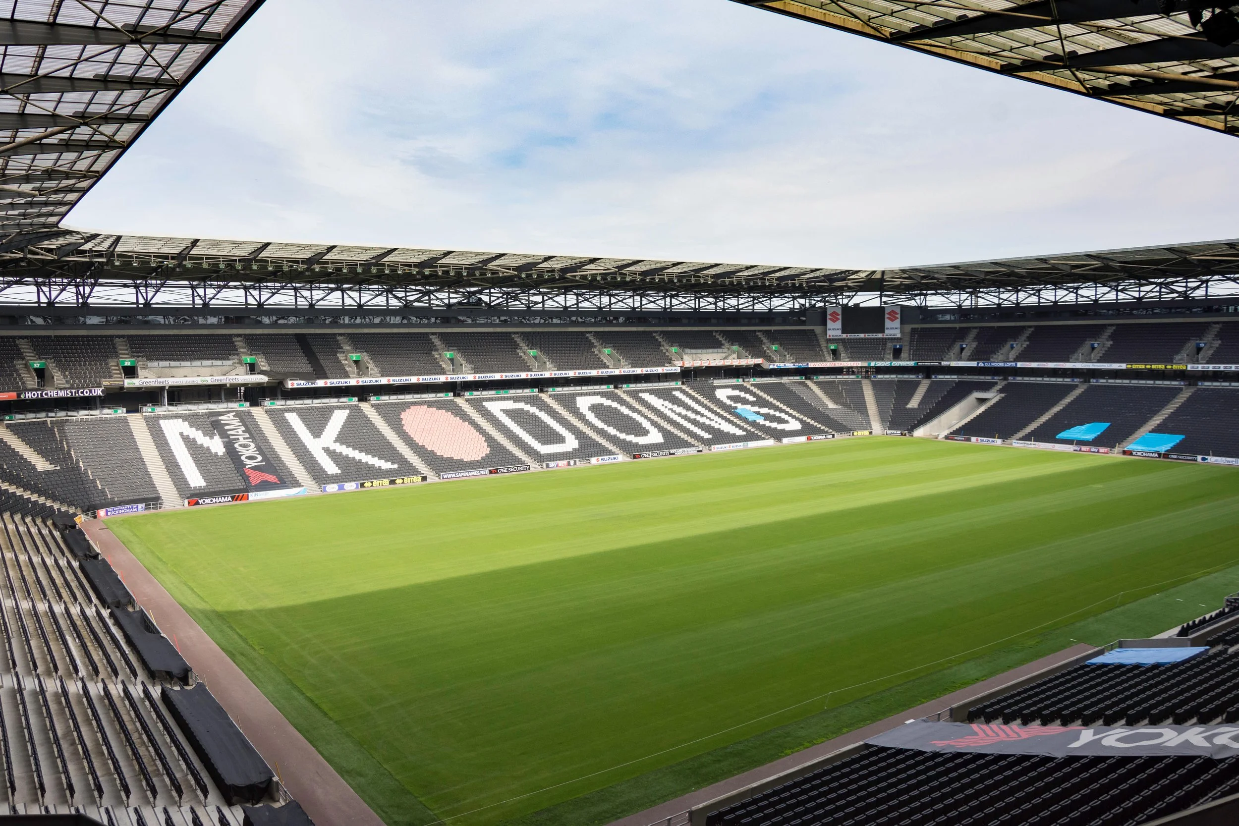 Empty soccer stadium with green field and seating area, large 'KODI' sign spelled out in seats, partially covered roof, and blue sky.