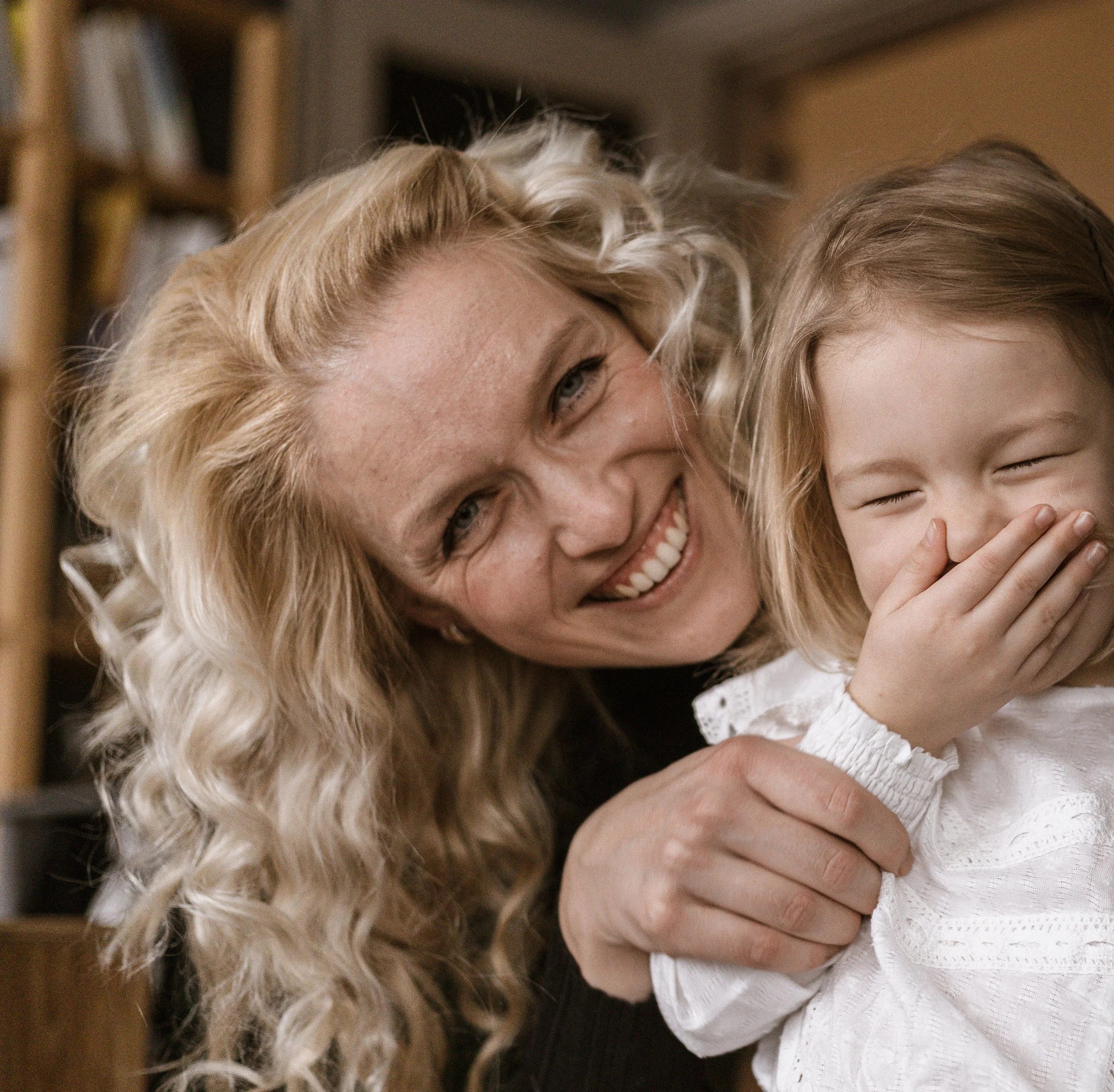 A woman with blonde, curly hair smiling and leaning close to a young girl with blonde hair. The girl is laughing with her eyes closed and hand covering her mouth, wearing a white top.