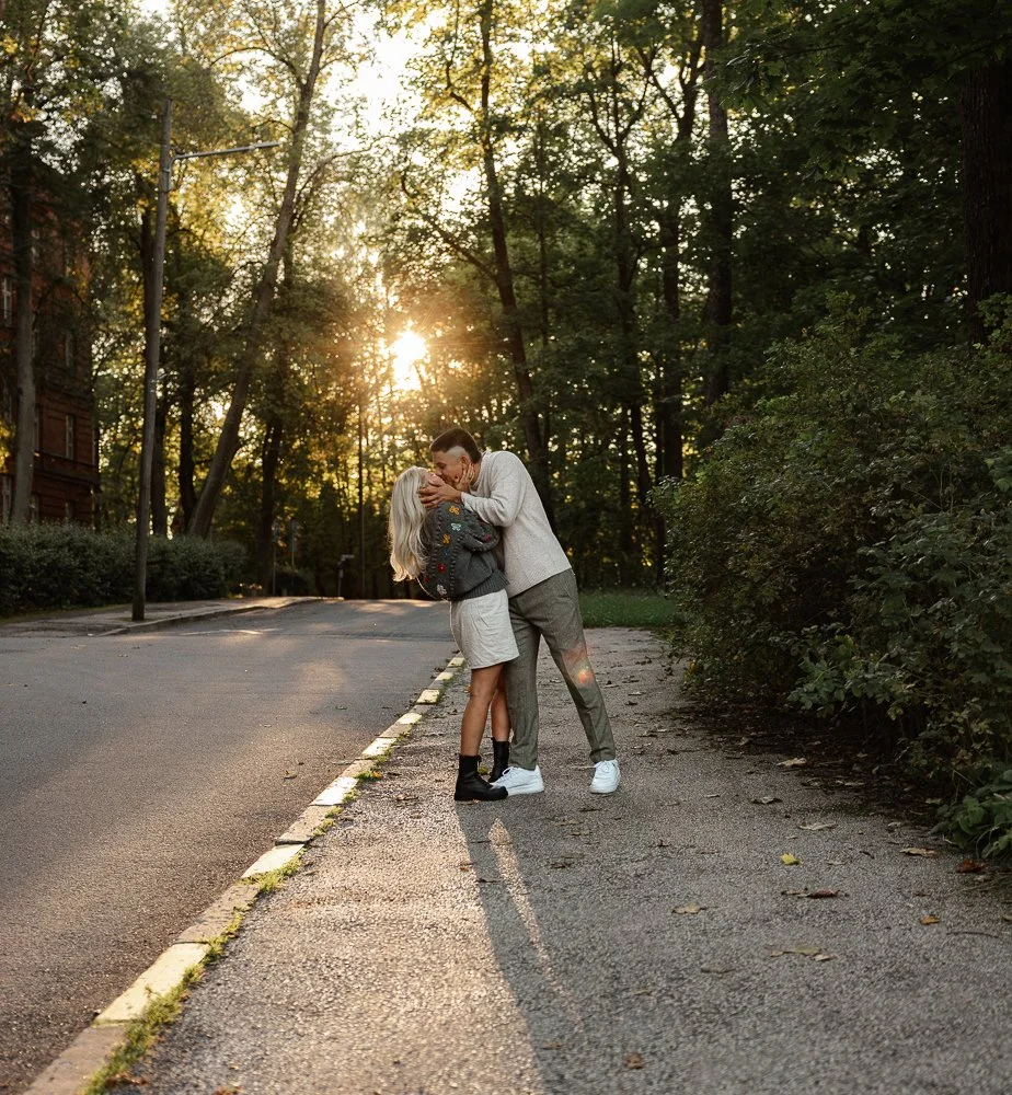 A couple is sharing a kiss on a sidewalk in a park during sunset, surrounded by trees and greenery.