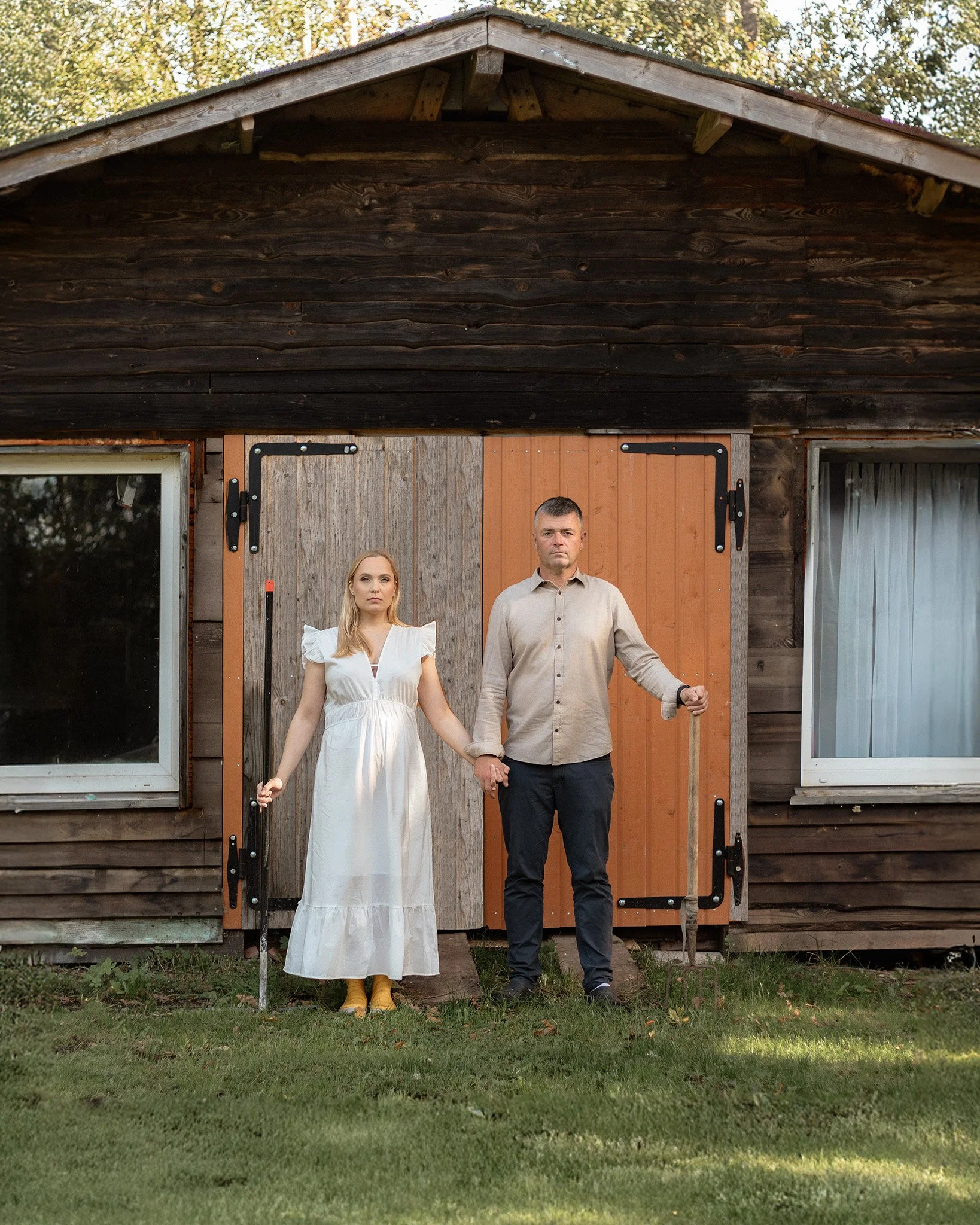 A man and woman holding hands stand in front of a rustic wooden barn with a sliding door. The woman is wearing a white dress and yellow boots, and the man is dressed in a beige shirt and dark pants.