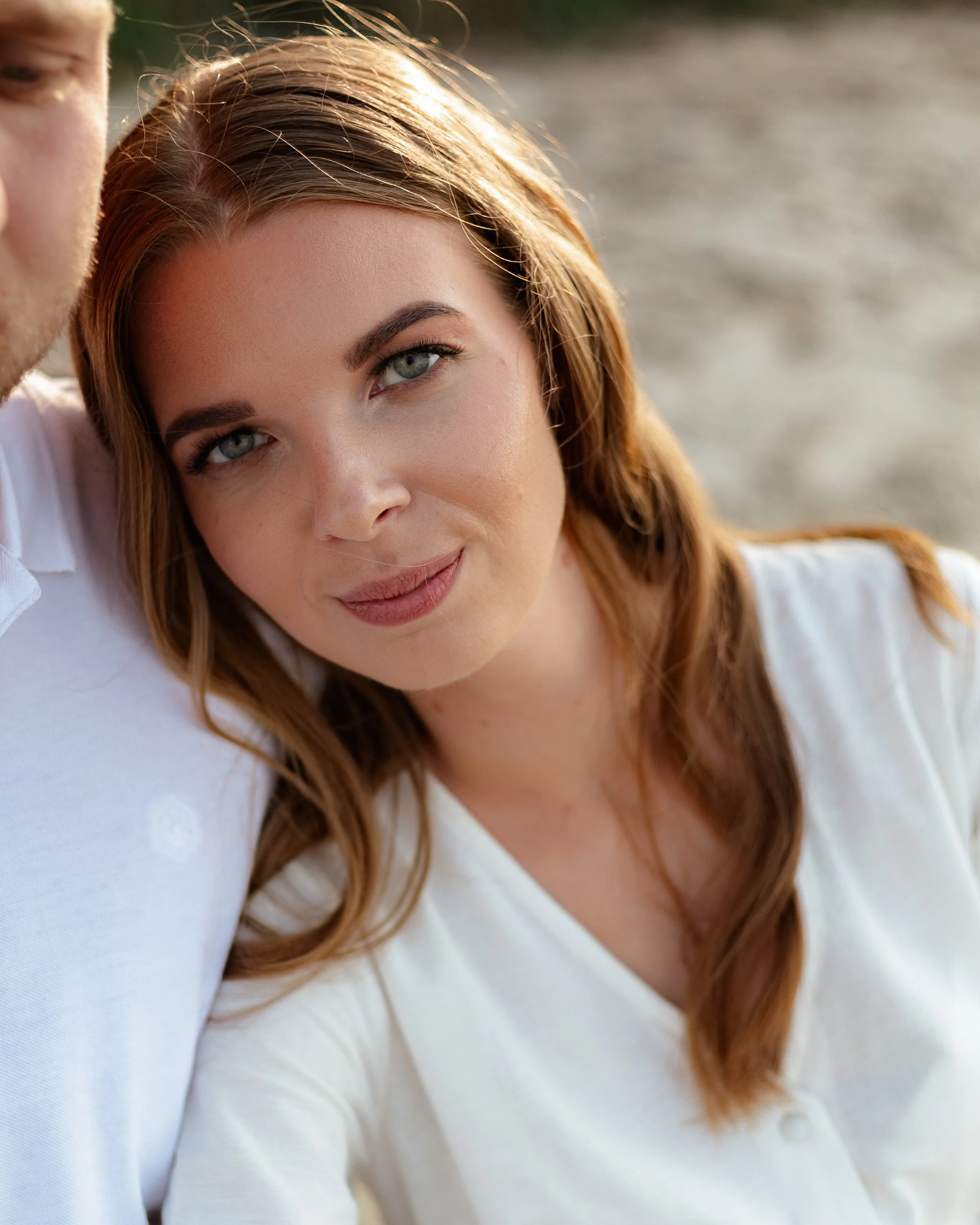A woman with red hair and blue eyes smiling close-up portrait, leaning on a man's shoulder, outdoors with blurred background.