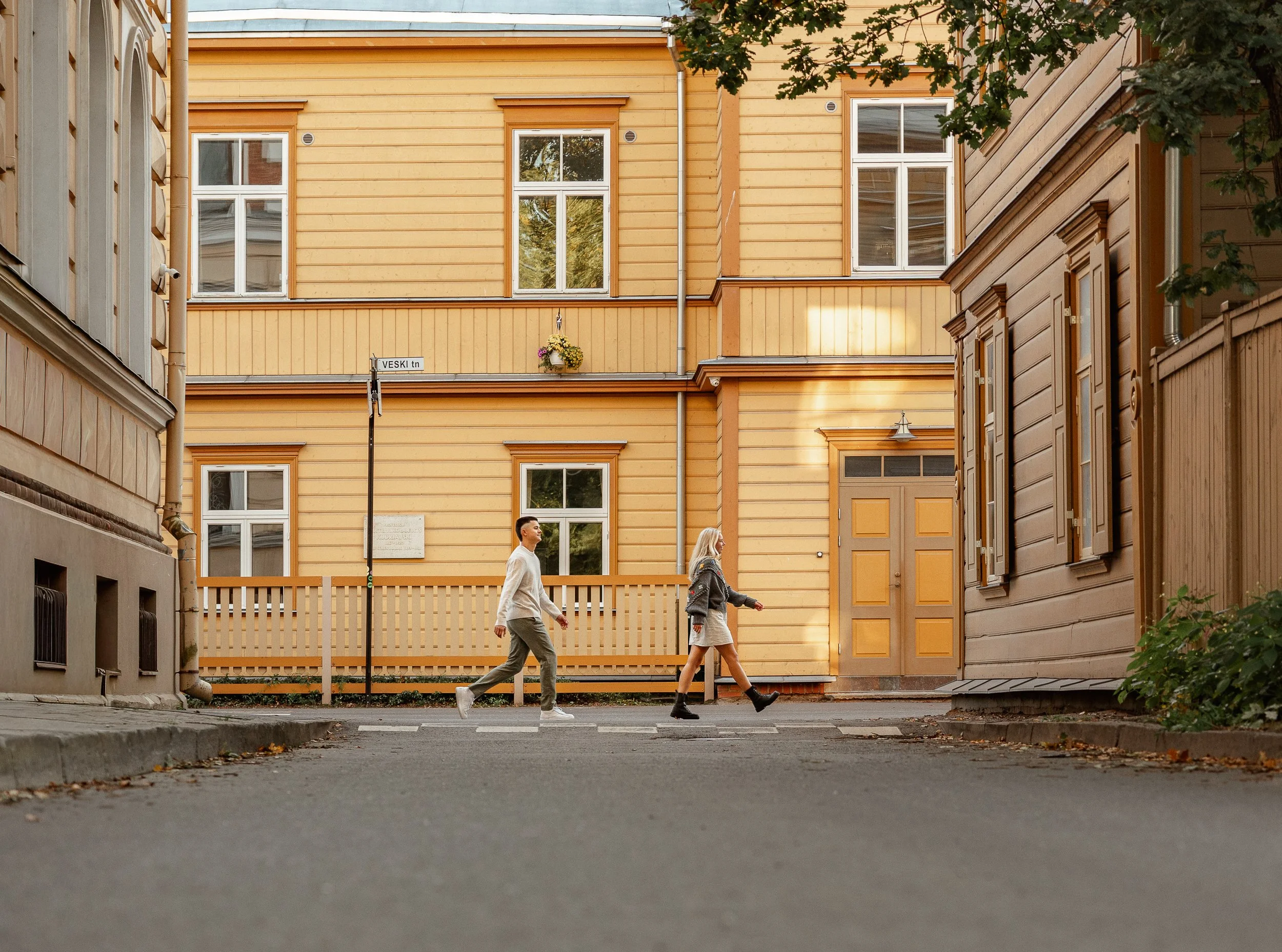 Two people walking across a street in front of yellow and brown wooden buildings with windows and a flower hanging from a pole.