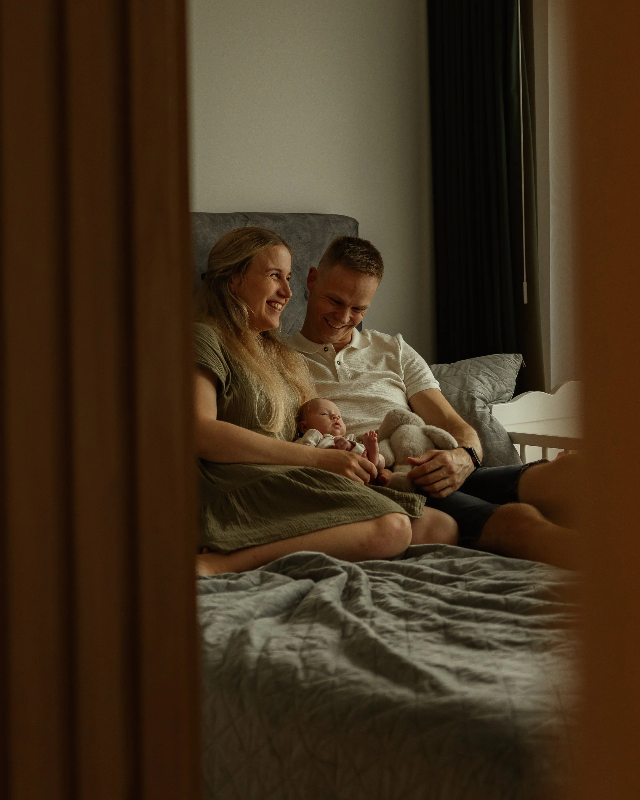 A family of three sitting on a bed, with a baby, smiling and looking at each other.