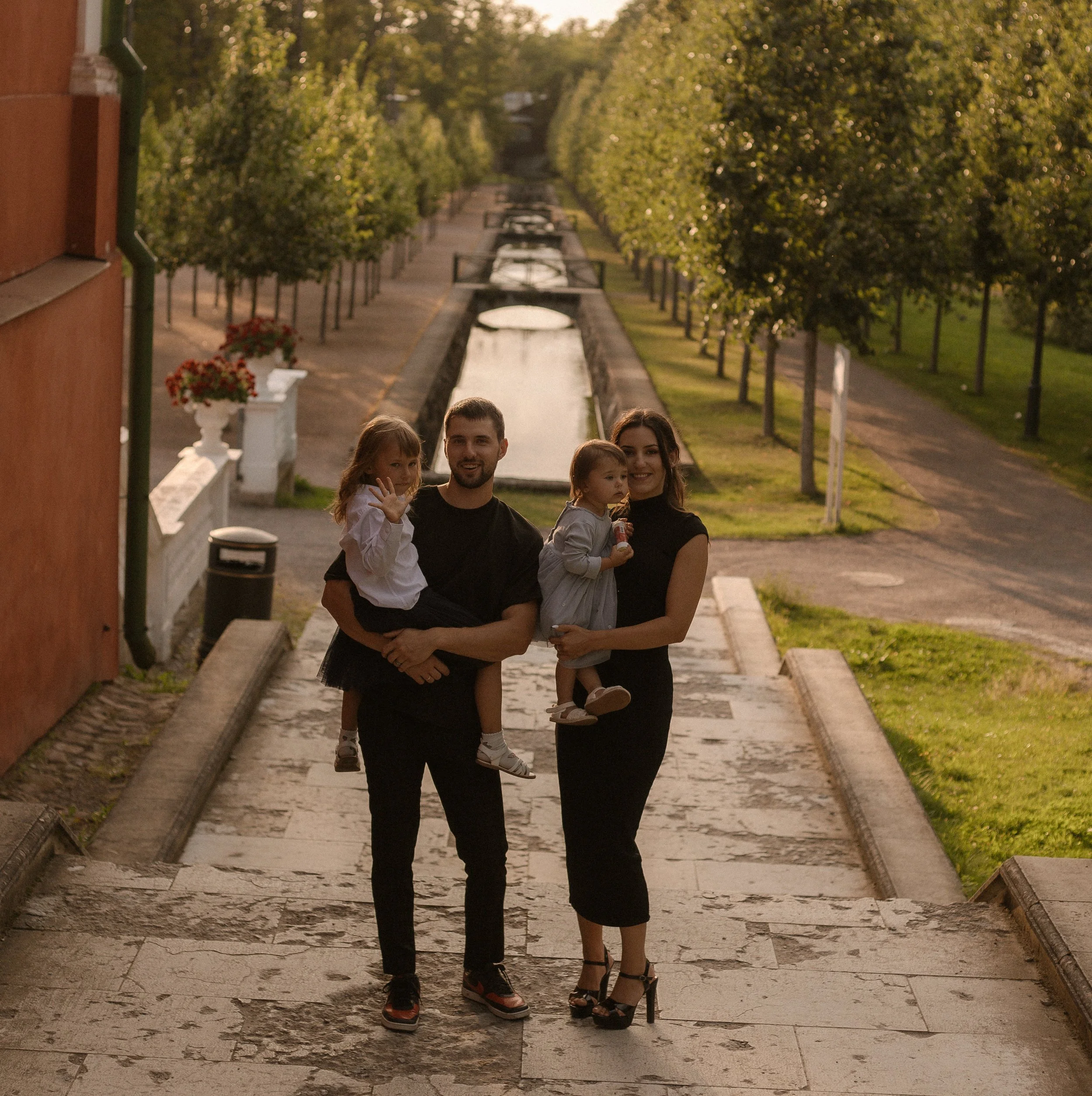 A family of four walking down a stone stairway near a canal, with trees on both sides and a bridge in the background, during sunset, smiling and enjoying the moment.