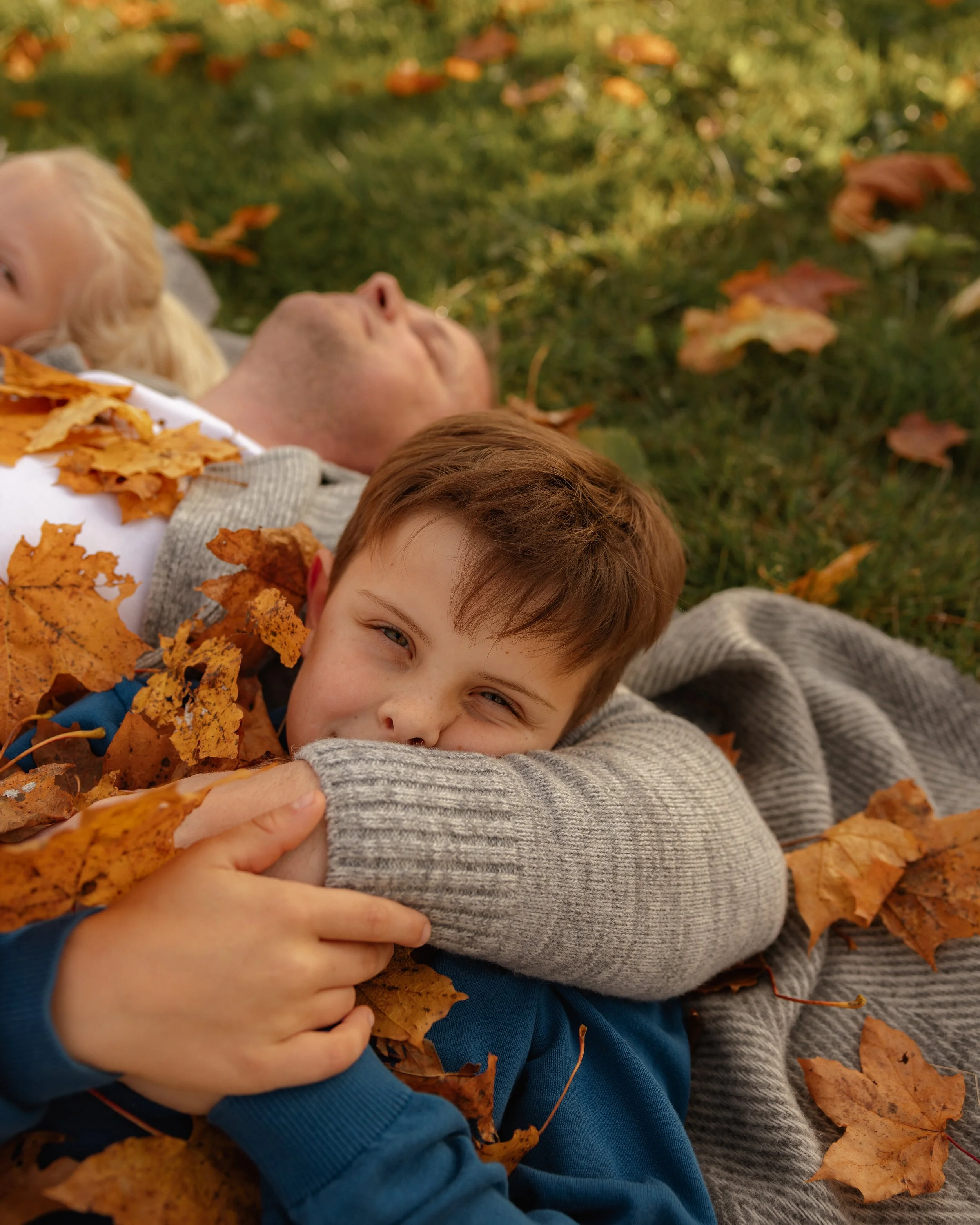 A boy lying on the grass with fallen autumn leaves, smiling and hugging a gray sweater sleeve, with two other people in the background also lying down surrounded by leaves.