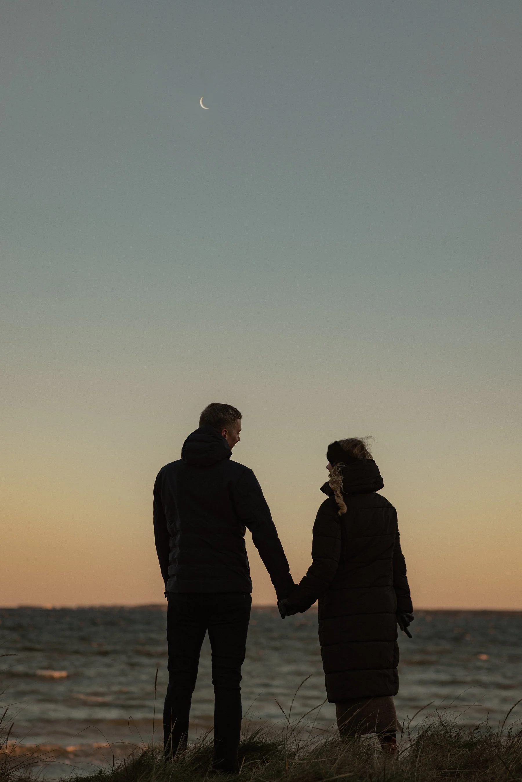 A couple standing on the beach during sunset, holding hands and looking at each other, with the moon visible in the sky.