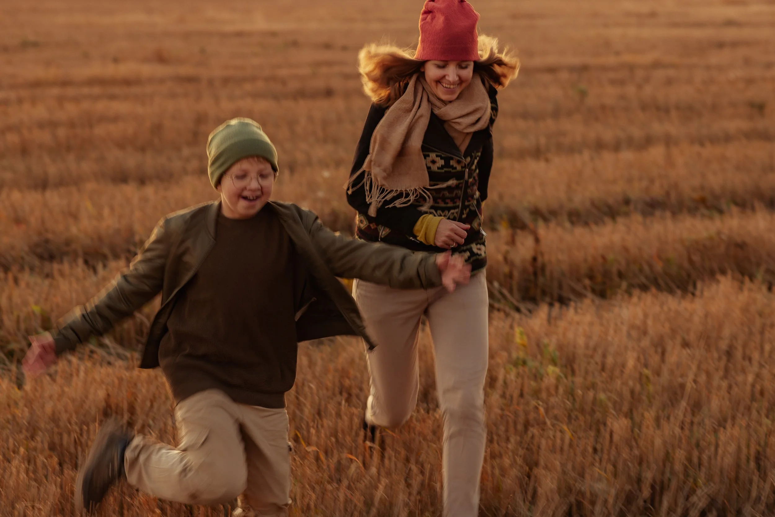 A woman and a young boy running through a field of tall, dry grass during sunset, smiling and holding hands.