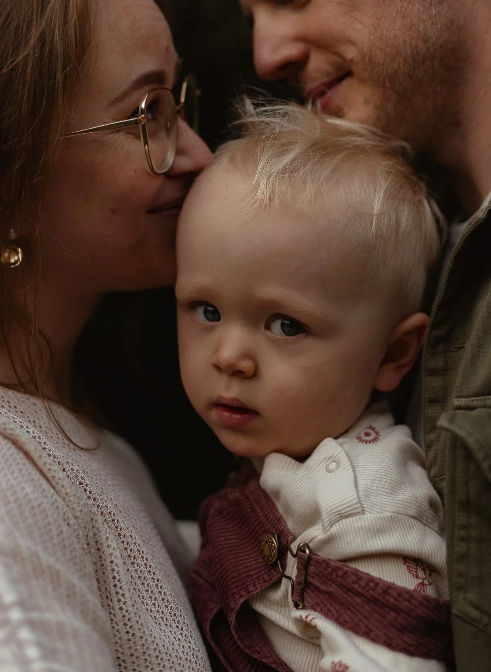 A family of three, including a woman with glasses, a man, and a young boy with blonde hair, close together. The woman and man are leaning in close to the child's face, who looks directly at the camera with a serious expression.
