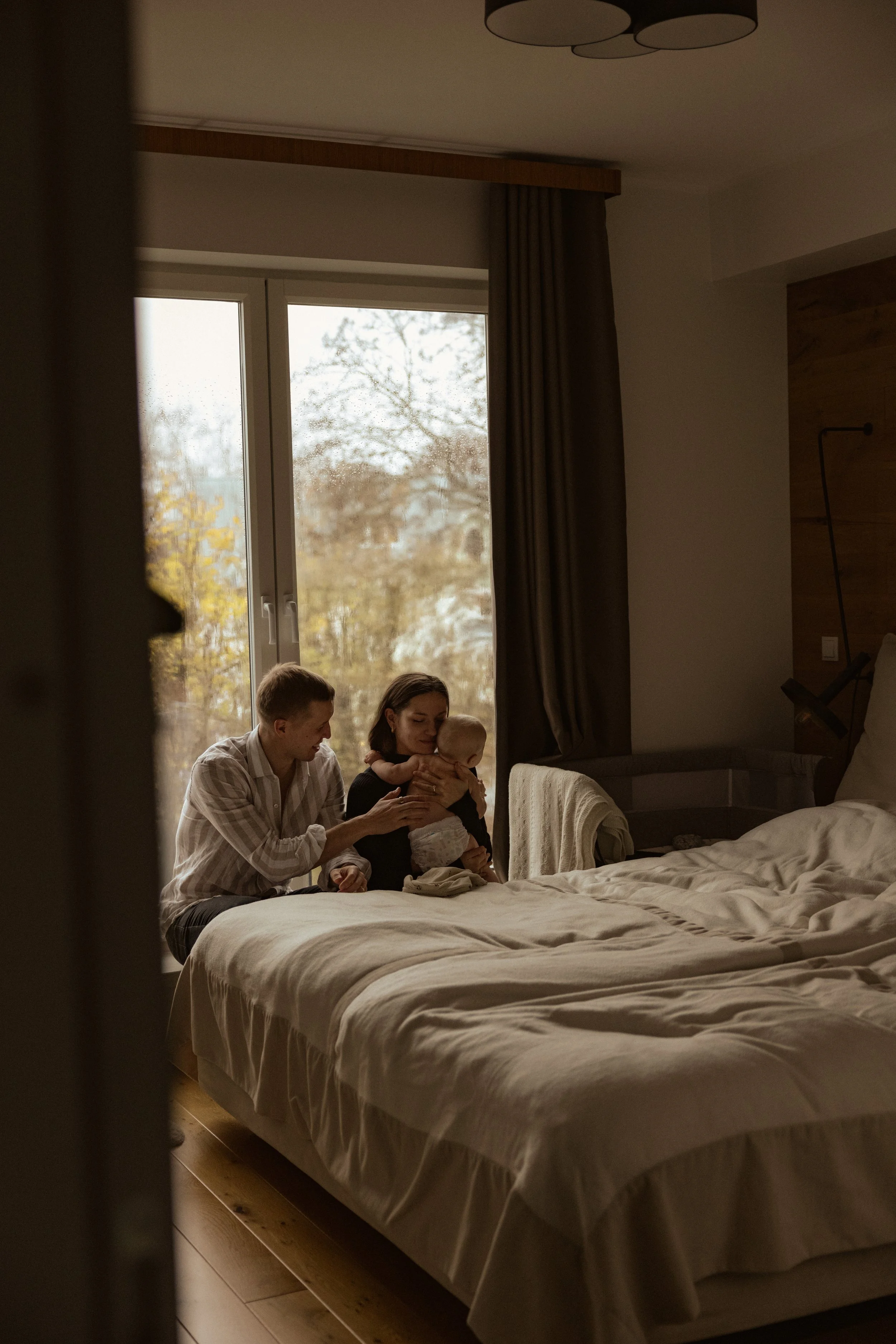 Family sitting on bed in a bedroom with large window showing trees outside, mother holding a baby, father reaching towards the baby.