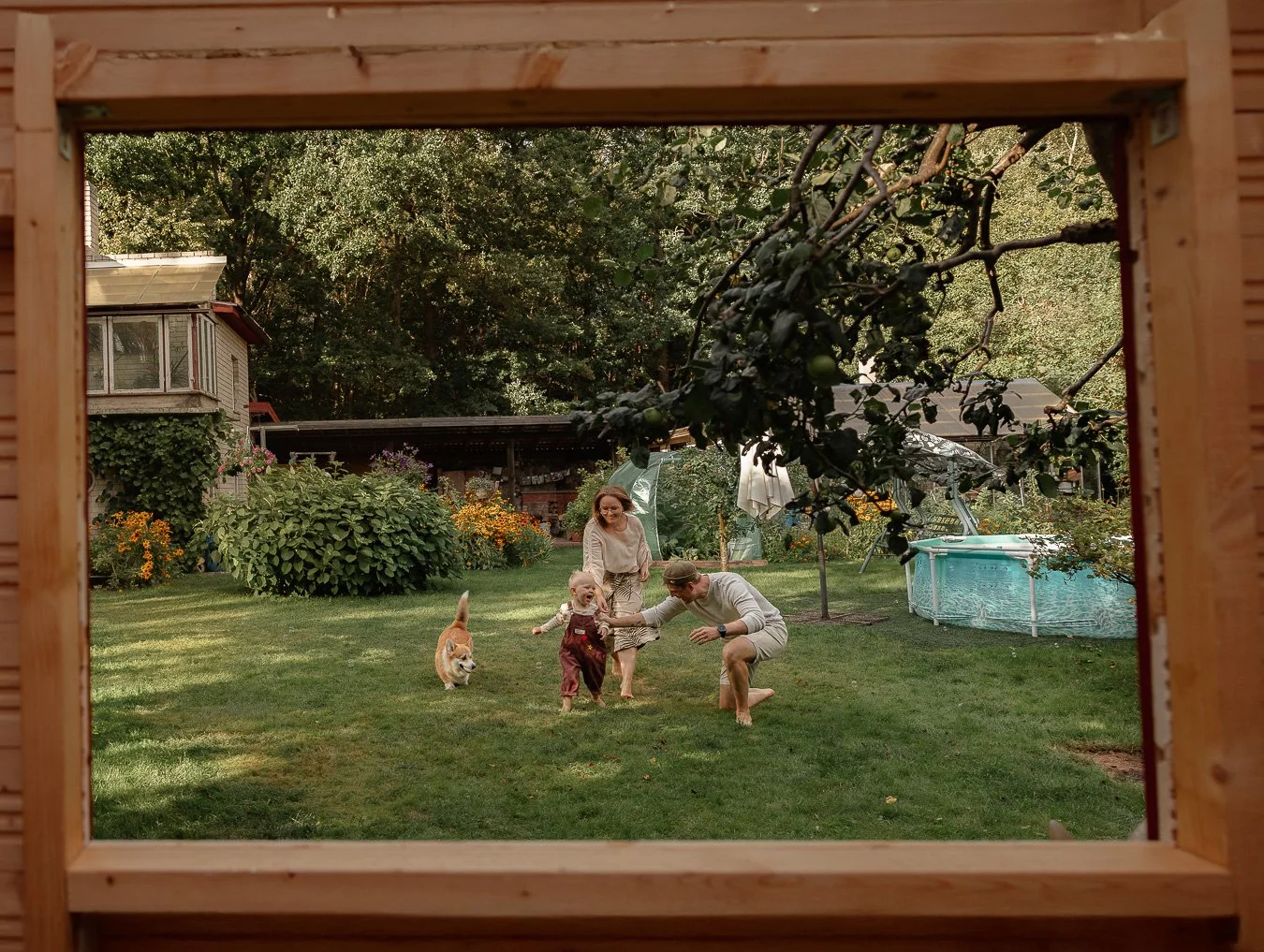 A family playing on the grass in a backyard, seen through a wooden frame, with trees, flowers, and a small pool in the background.