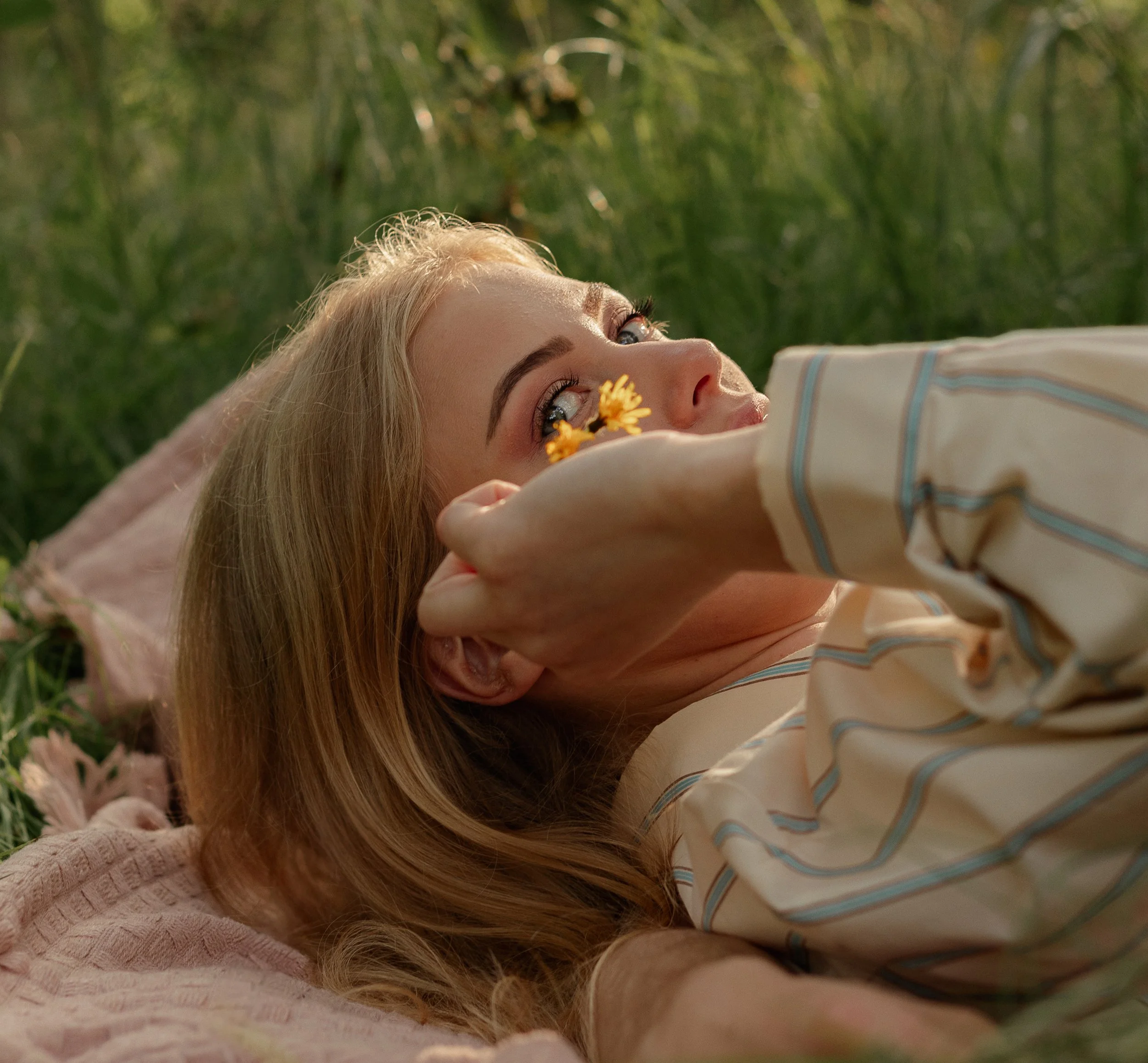 A woman with blond hair lying on her back in a grassy field, holding a yellow flower near her face, with her head resting on a pink blanket, and looking upward.
