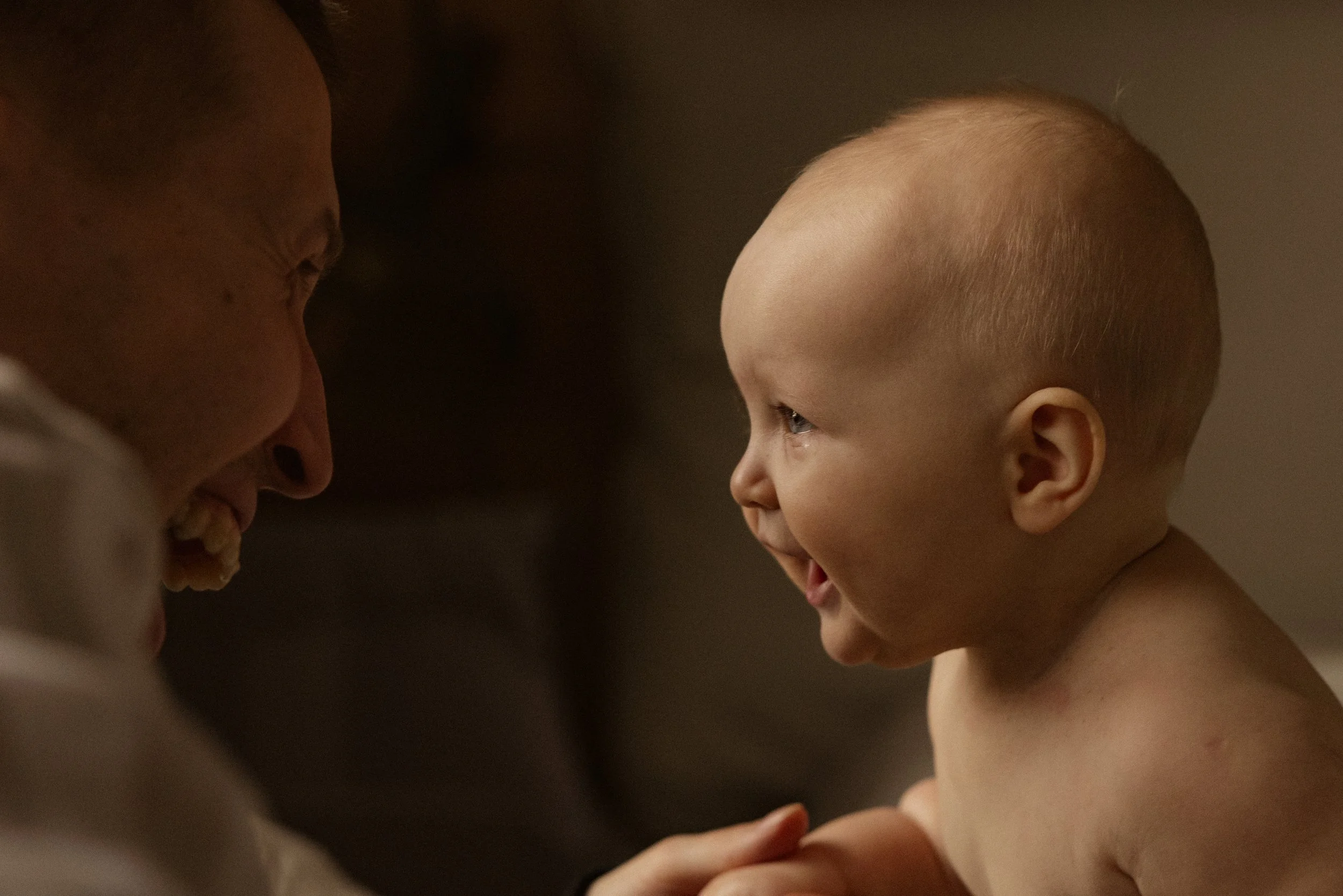 A young child with a shaved head smiling and laughing while facing an adult, both sharing a joyful moment.