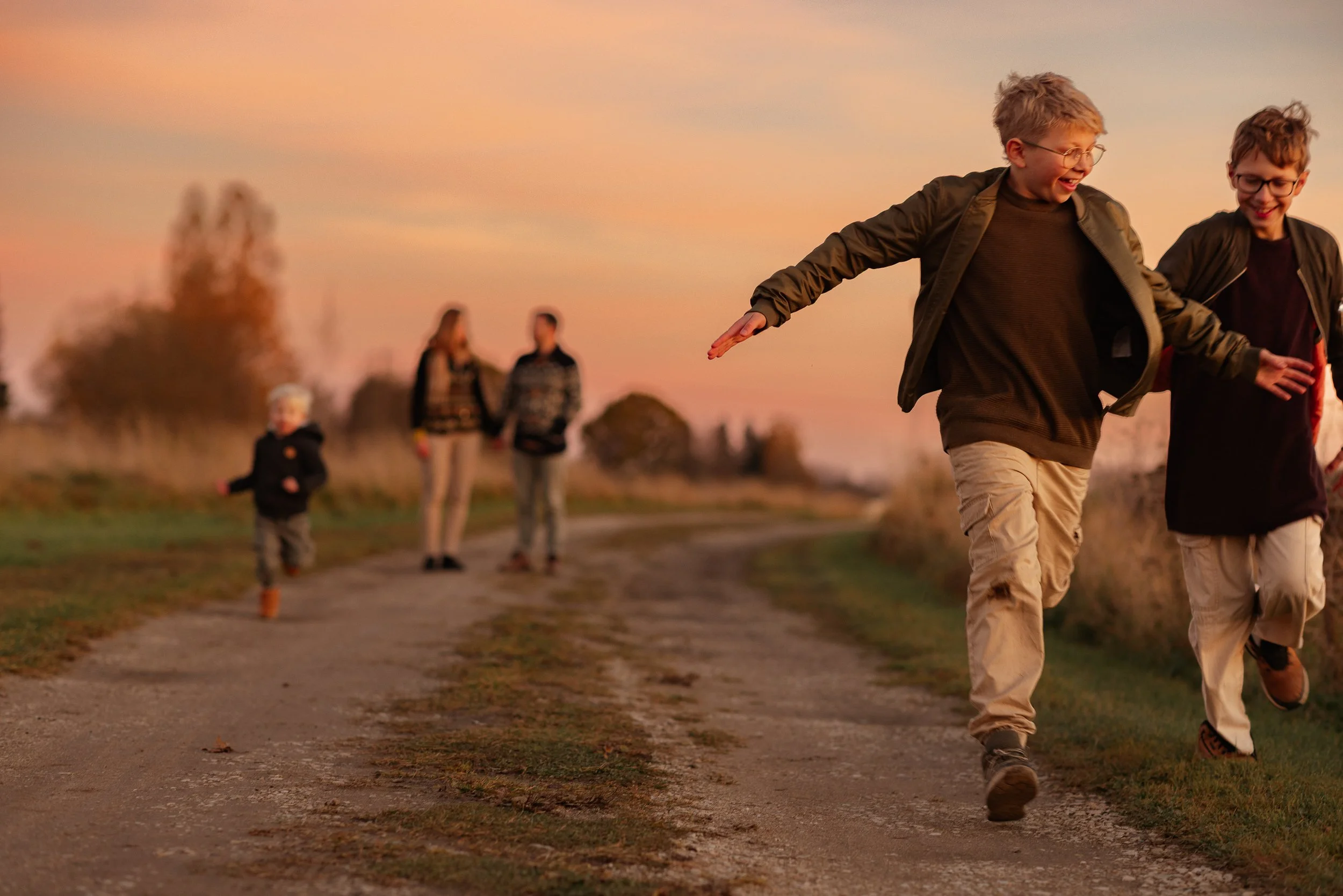 A group of five people, including two children and two adults, enjoying a walk on a dirt path during sunset. The children are running and laughing, while the adults walk behind. The sky is colorful with orange and pink hues.