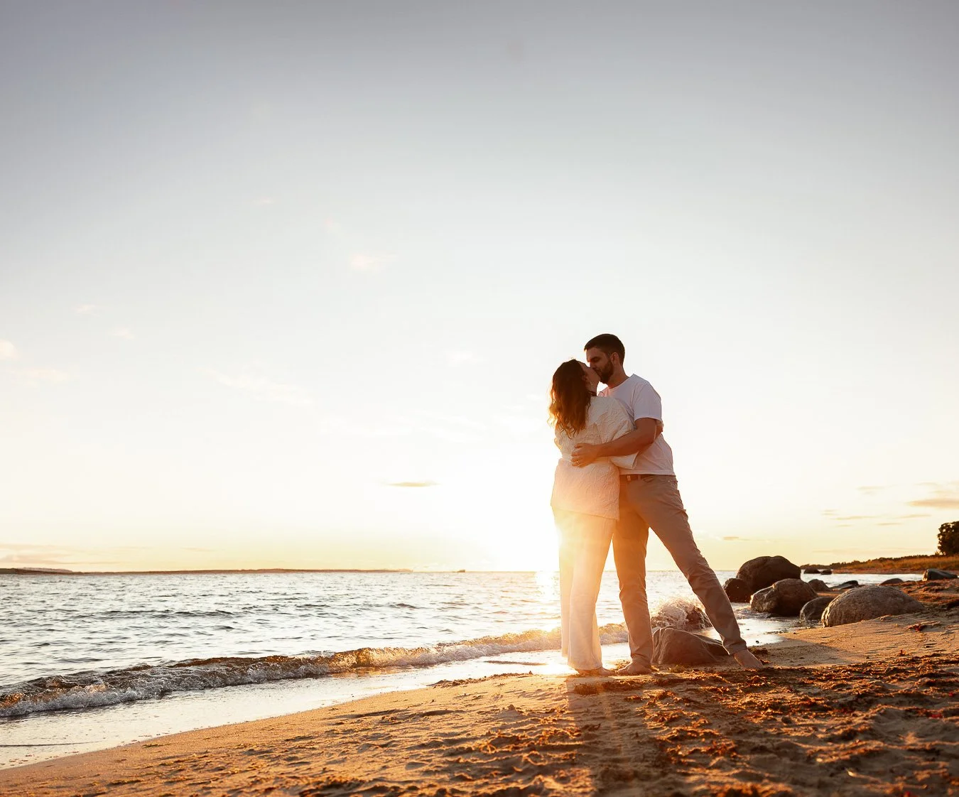 A couple embraces on a beach at sunset, with rocks in the background and the sun near the horizon.