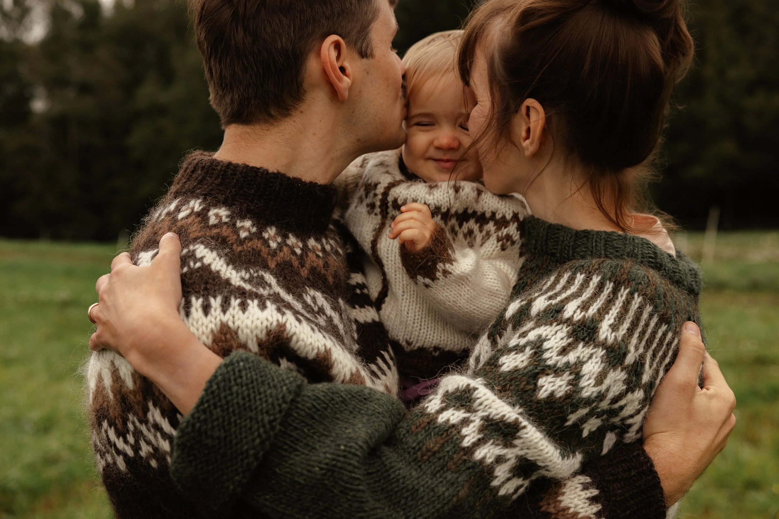 A family of three embracing outdoors in autumn; the mother and father are kissing the child's cheek, who is smiling.