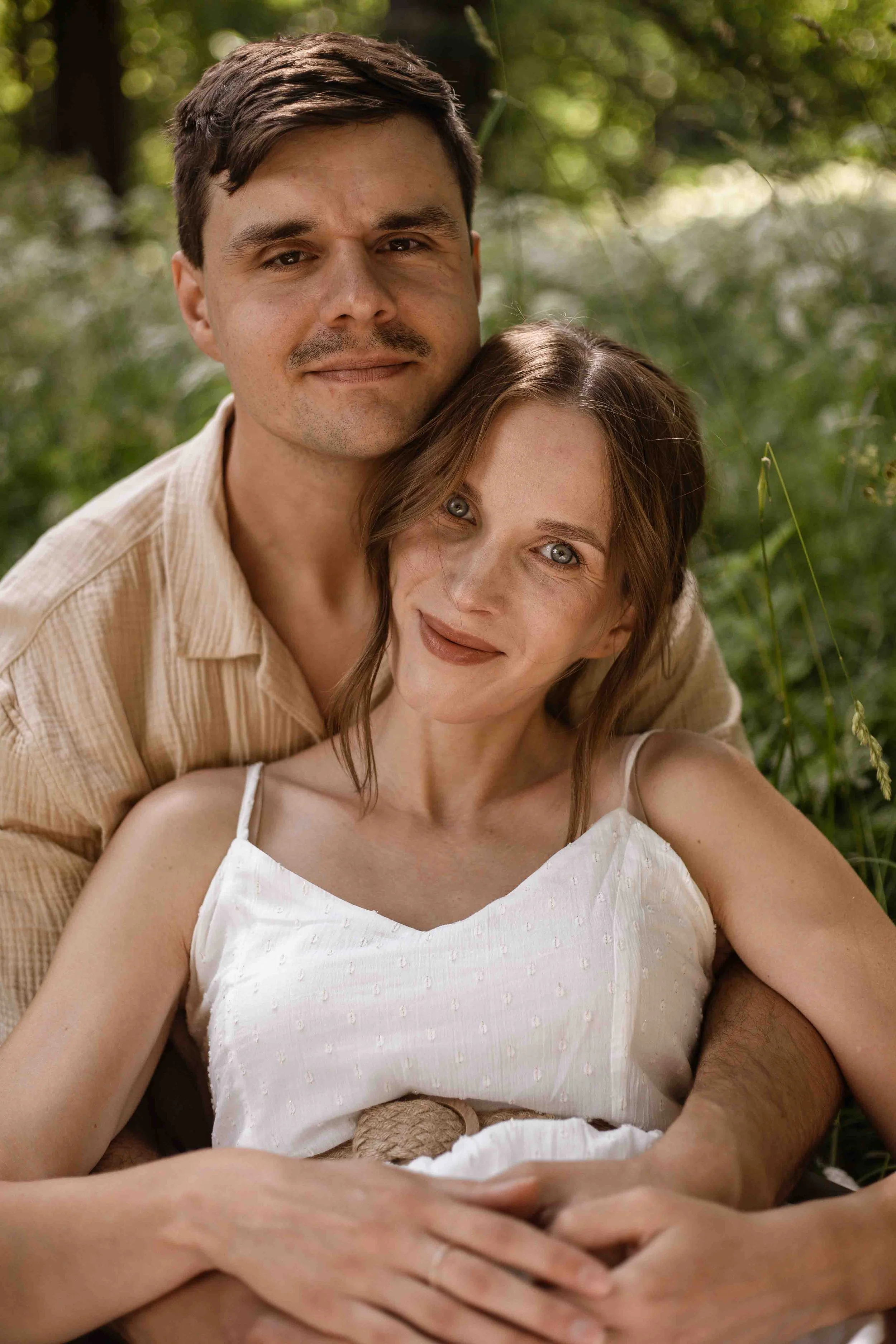 A man and woman sitting outdoors surrounded by greenery. The man has short dark hair and a mustache, and is wearing a beige shirt. The woman has shoulder-length reddish-brown hair, blue eyes, and is wearing a white tank top. They are smiling gently and appear to be happy together.