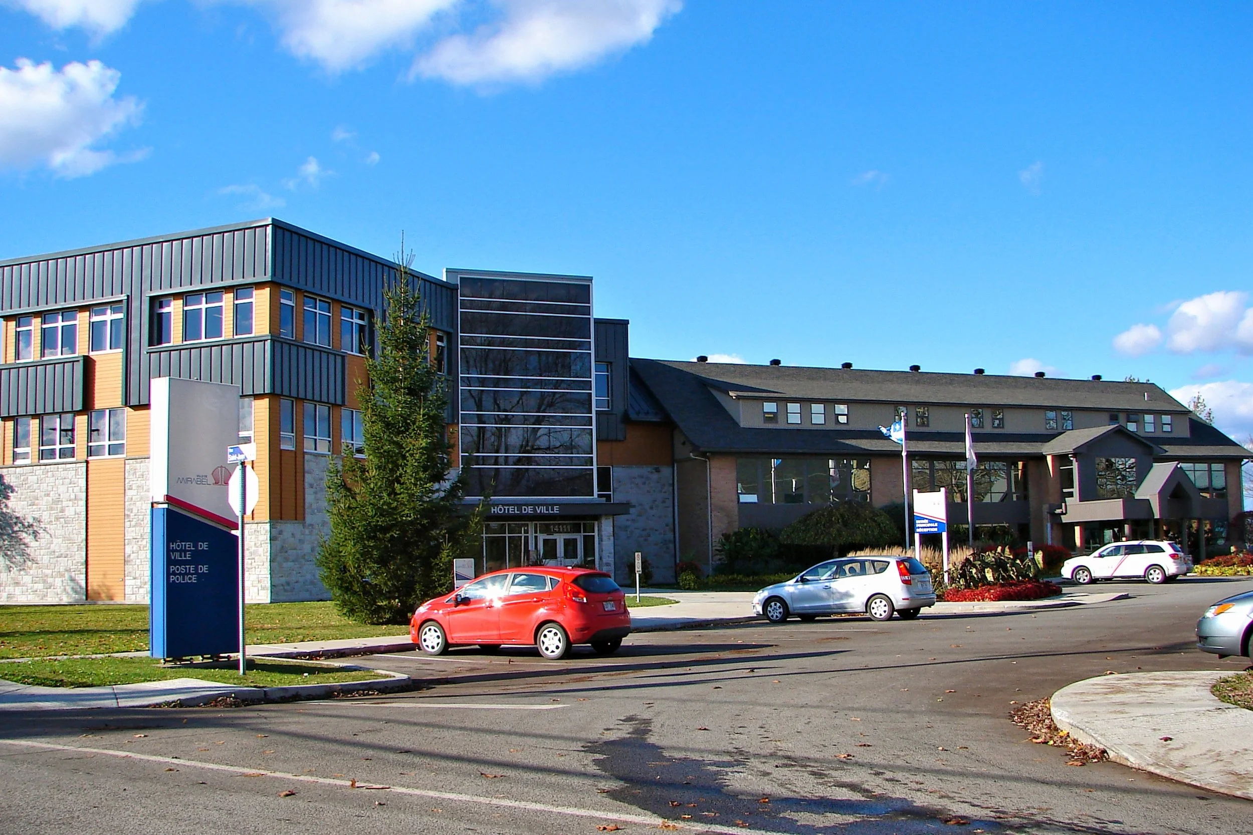 Bâtiment moderne avec plusieurs étages, fenêtres, et un parking avec des voitures, sous un ciel bleu avec quelques nuages.
