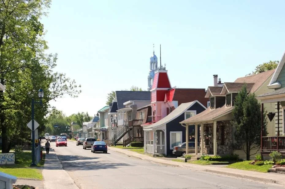Une rue résidentielle avec des maisons colorées, des voitures garées et des arbres verts sous un ciel dégagé.