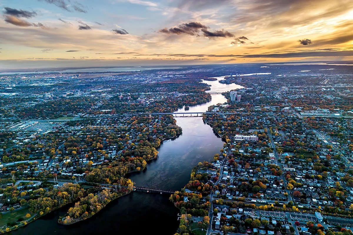 Vue aérienne d'une ville avec un fleuve qui serpente au milieu, entourée d'arbres aux couleurs automnales, sous un ciel orageux avec des nuages et un coucher de soleil.