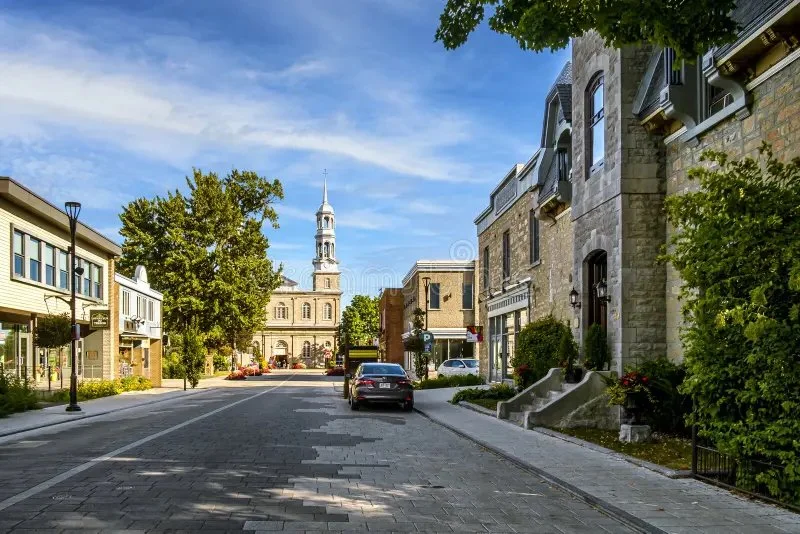 Une rue calme dans une ville avec des bâtiments résidentiels en pierre et une église au centre avec une tour et un clocher, sous un ciel bleu avec quelques nuages, arbres verdoyants le long de la rue, et des voitures stationnées.