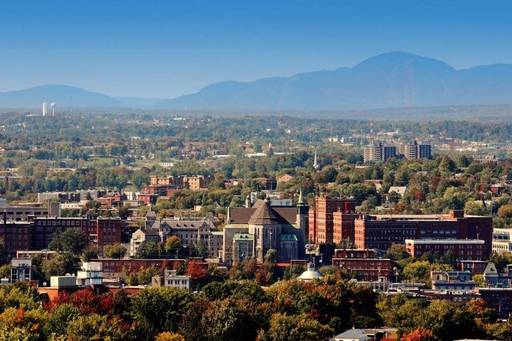 Vue panoramique d'une ville avec des bâtiments historiques, des arbres et des montagnes à l'horizon lors d'une journée ensoleillée.