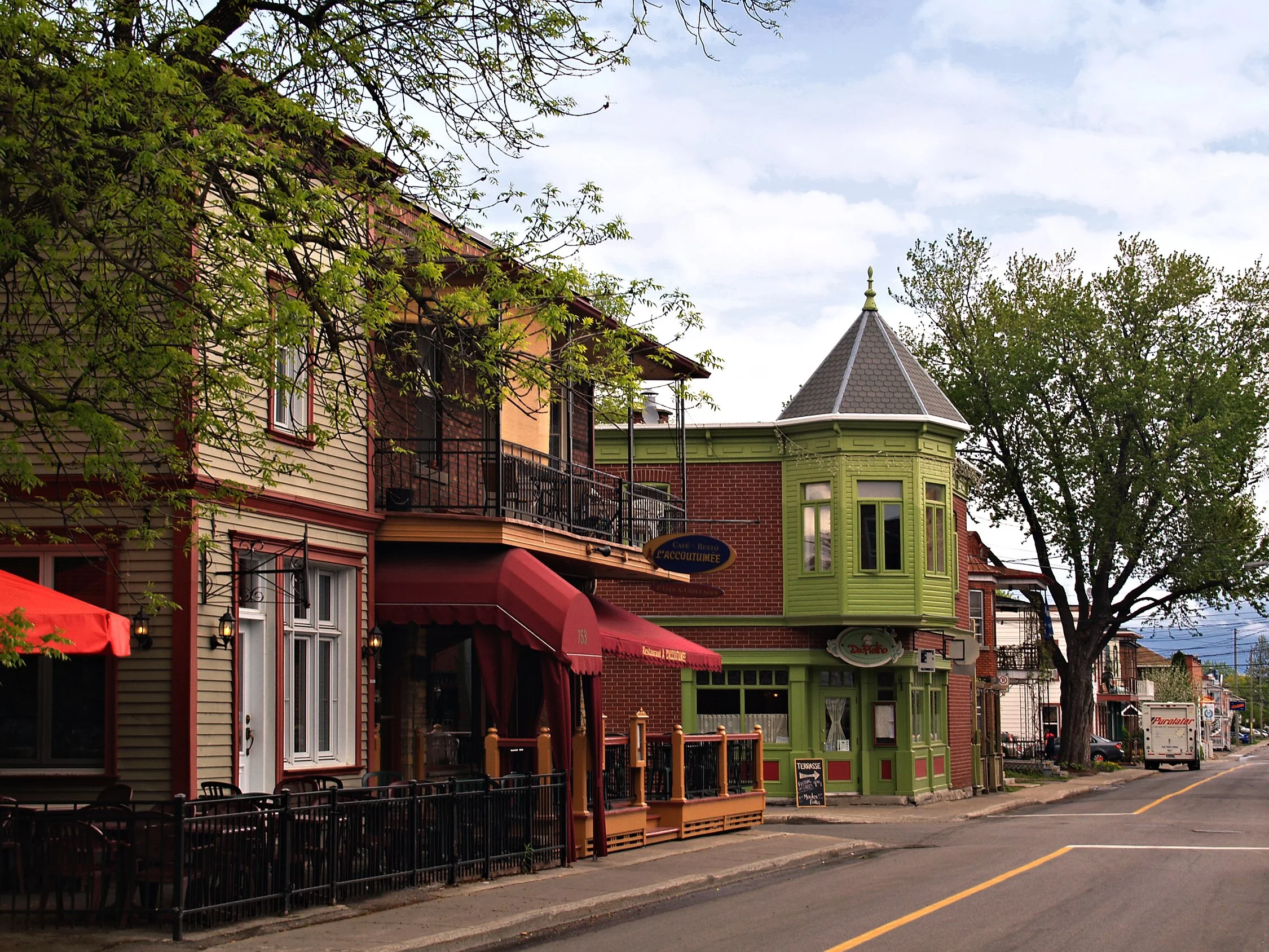 Rue commerçante avec des bâtiments colorés dont un vert, coiffés de toits en pente, et des terrasses extérieures. Il y a des arbres avec des feuilles vertes et un ciel partiellement nuageux.