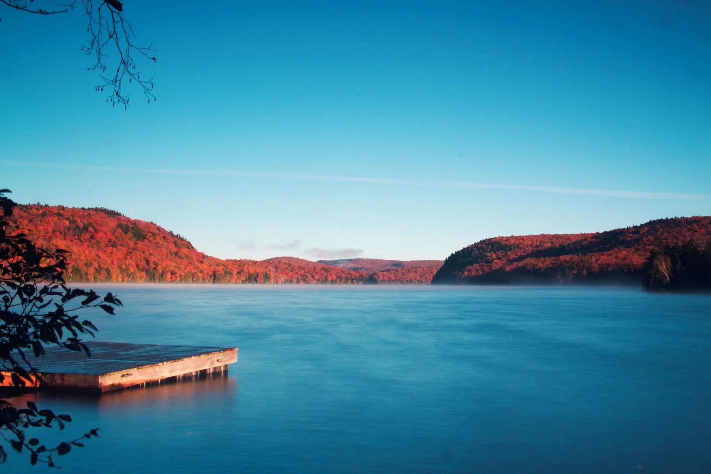 Lac entouré de collines boisées aux couleurs automnales, un quai en bois au premier plan, ciel clair et bleu.